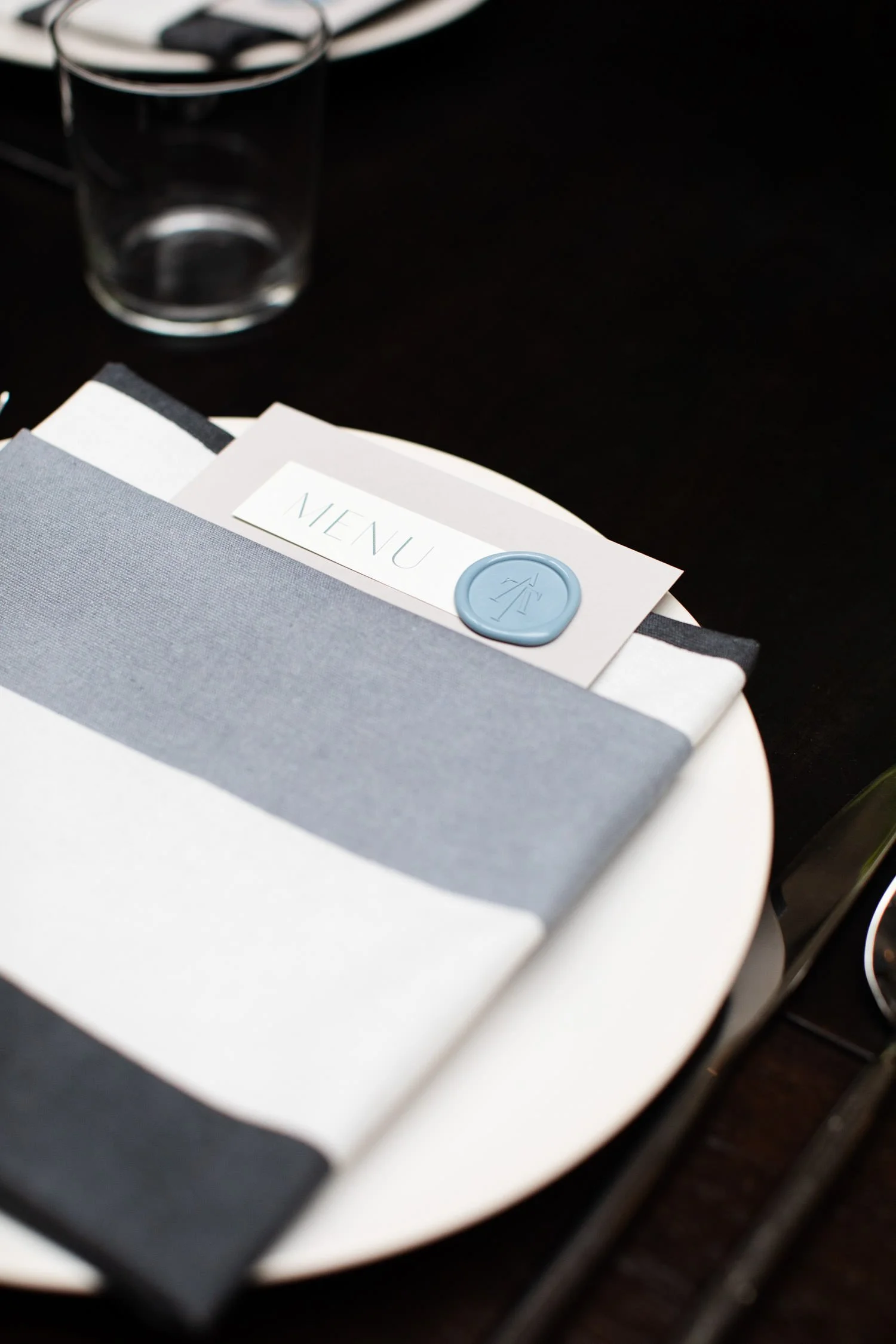 Black long tables and chairs with white tableware and custom-designed menu stationary and wax initials sealing it.. White daisies are placed perfectly. Minneapolis rehearsal dinner, Spoon and Stable restaurant, Designed by Rocket Science Events.
