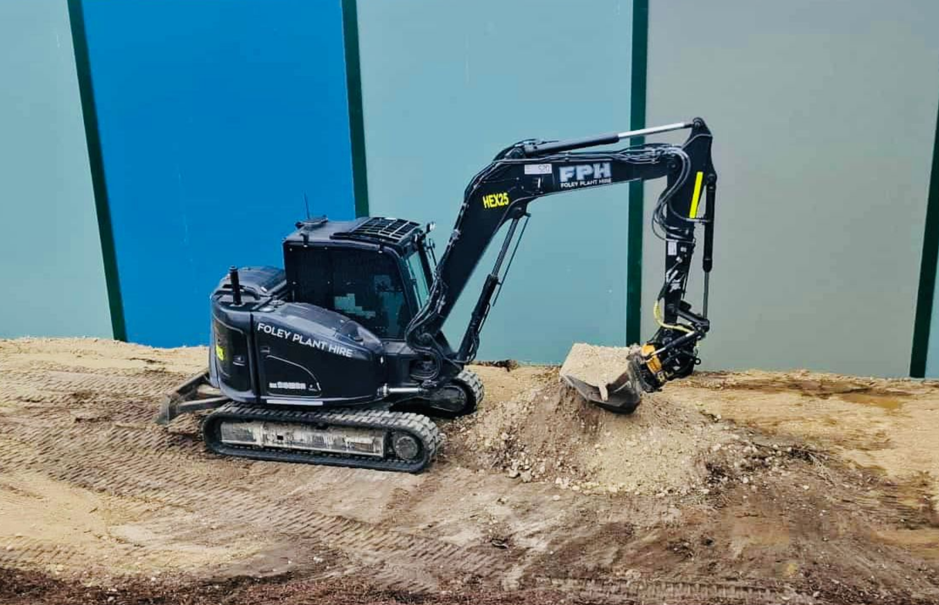 A small black excavator with the words 'Foley Plant Hire' on its side is digging into a mound of dirt on a construction site, with a green and blue wall behind it.