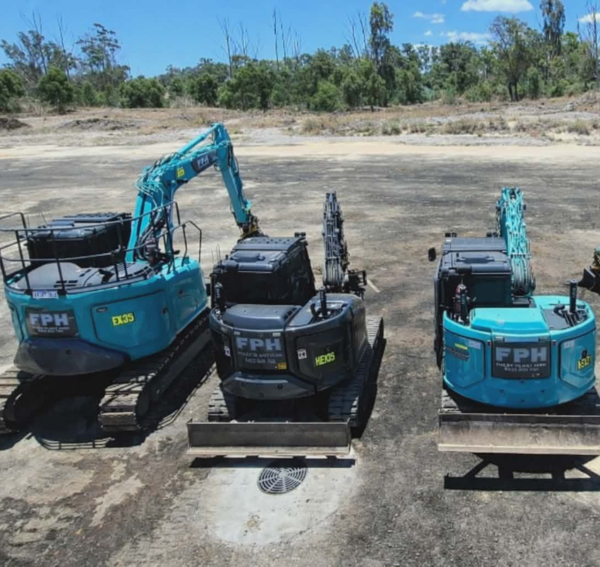 Three mini excavators on a gravel surface with a rural background, blue sky, and trees.