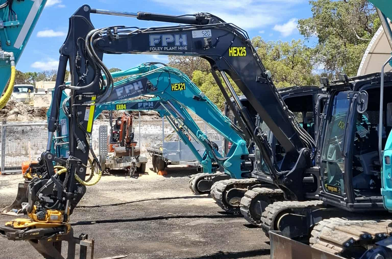 Two Robotic excavators at a construction site, the one in the foreground is black with yellow accents, and the one in the background is teal, both labeled with model and company branding.