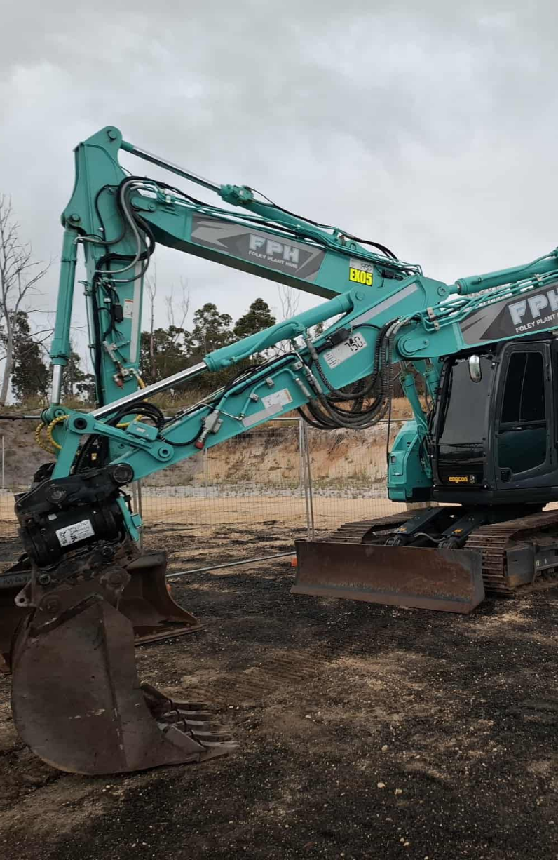 A teal-colored excavator with the brand name FPH on the arm, sitting on a construction site with a dirt ground and fenced perimeter. Trees and cloudy sky in the background.