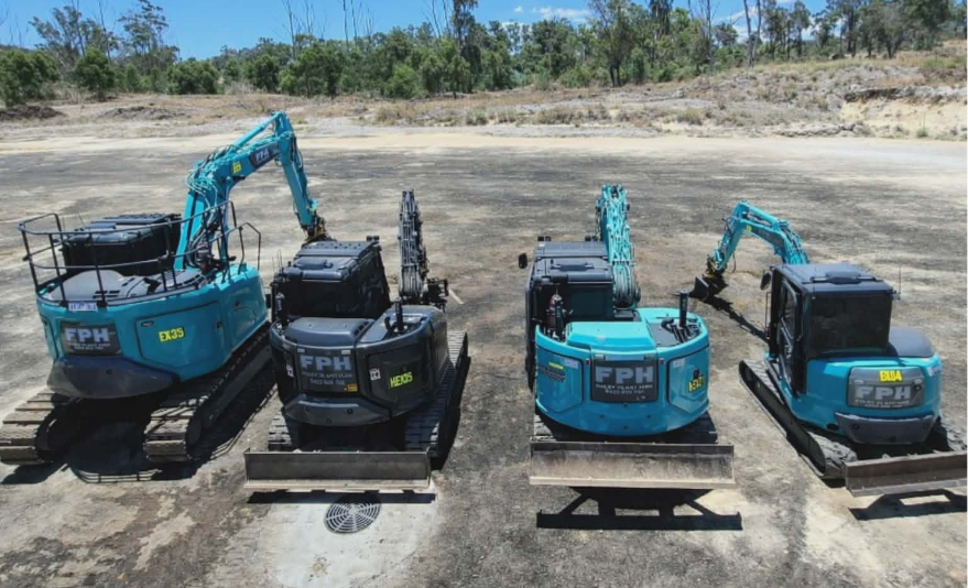 Four small excavators parked on a dirt surface with a tree line and clear sky in the background.