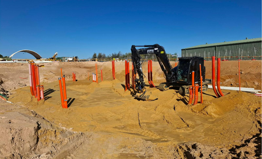 Construction site with an excavator working on underground pipe installation, orange safety markers, and a clear blue sky.