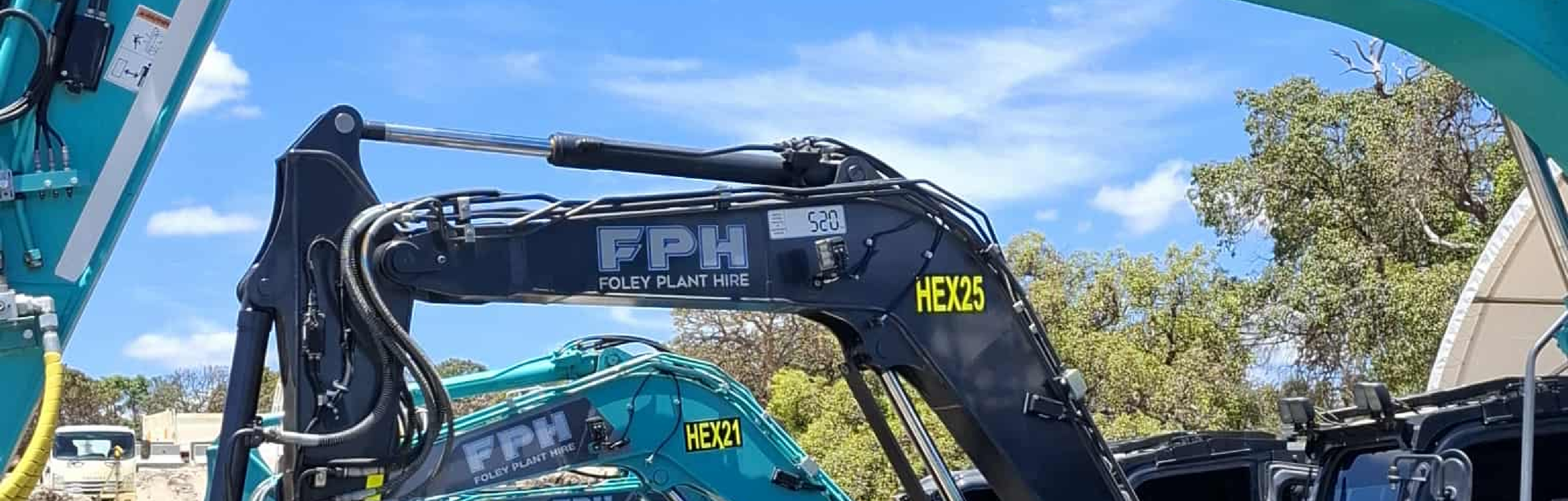 Close-up of a black hydraulic excavator arm with blue and yellow labels, with clear sky and trees in the background.