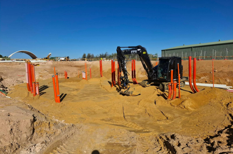 Construction site with excavator and orange pipes in sandy ground under a clear blue sky.
