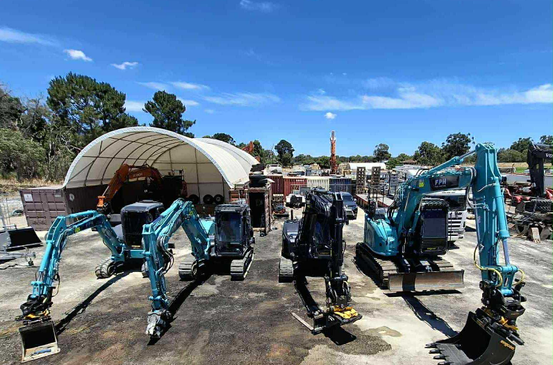 Three blue miniature excavators lined up on a paved lot with a curved white storage tent and trees in the background.
