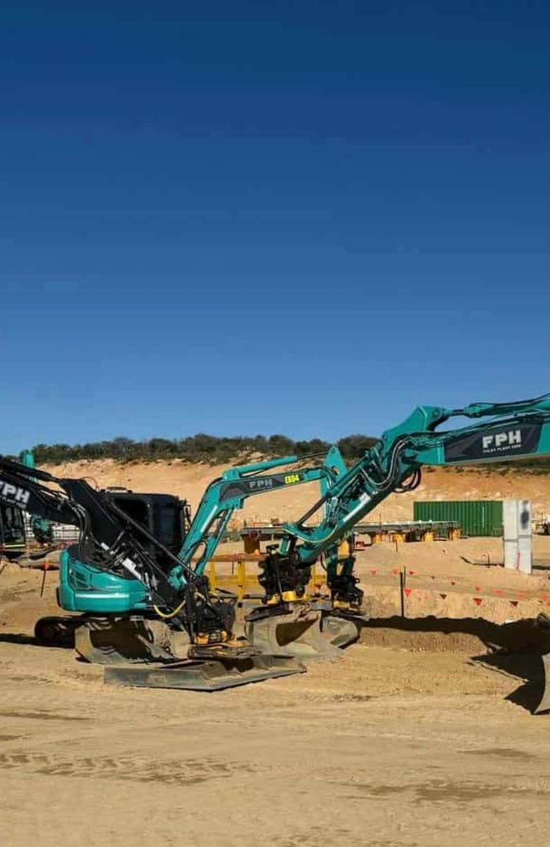 Construction site with two teal-colored excavators on sandy ground, backed by a small hill and a clear blue sky, with orange safety flags and containers in the background.