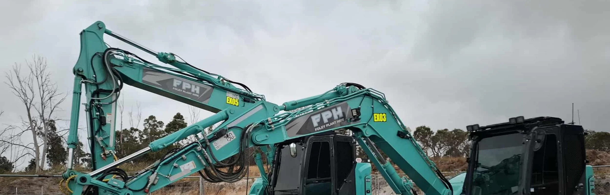 A large teal-colored excavator with hydraulic arms and parts labeled with logos and yellow tags, positioned outdoors under a cloudy sky with sparse trees in the background.