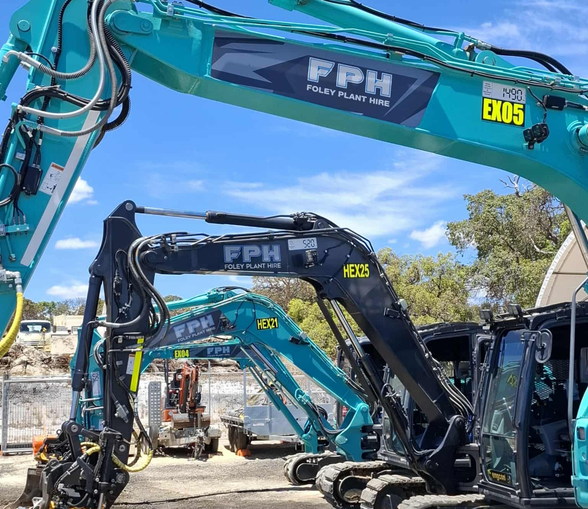 Two blue excavators with black and yellow labels on their arms, parked outdoors on a construction site with trees and other construction equipment in the background, and a clear blue sky overhead.
