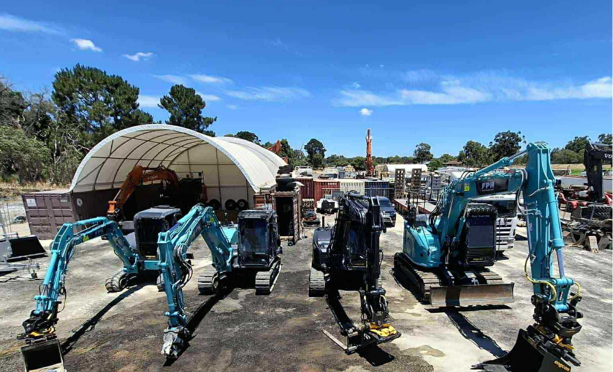 Multiple compact excavators lined up outdoors on a paved surface at a construction site, with a large curved tent and various equipment and machinery in the background under a blue sky.