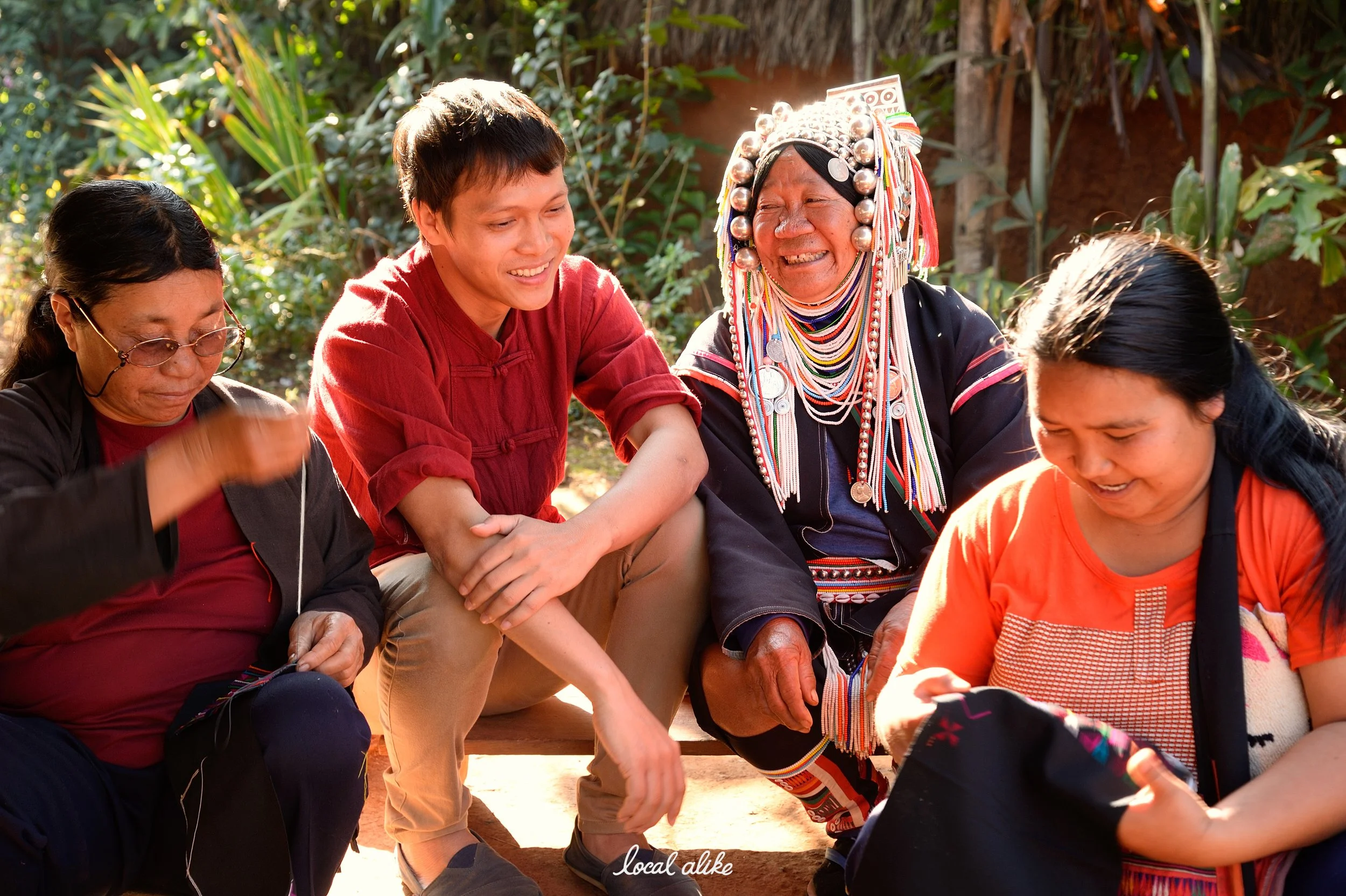 Four people sitting outdoors, smiling and laughing. They include women dressed in traditional and modern clothing, with one woman wearing traditional elaborate jewelry and headgear. The setting appears to be in a lush, green area with sunlight filtering through the trees.