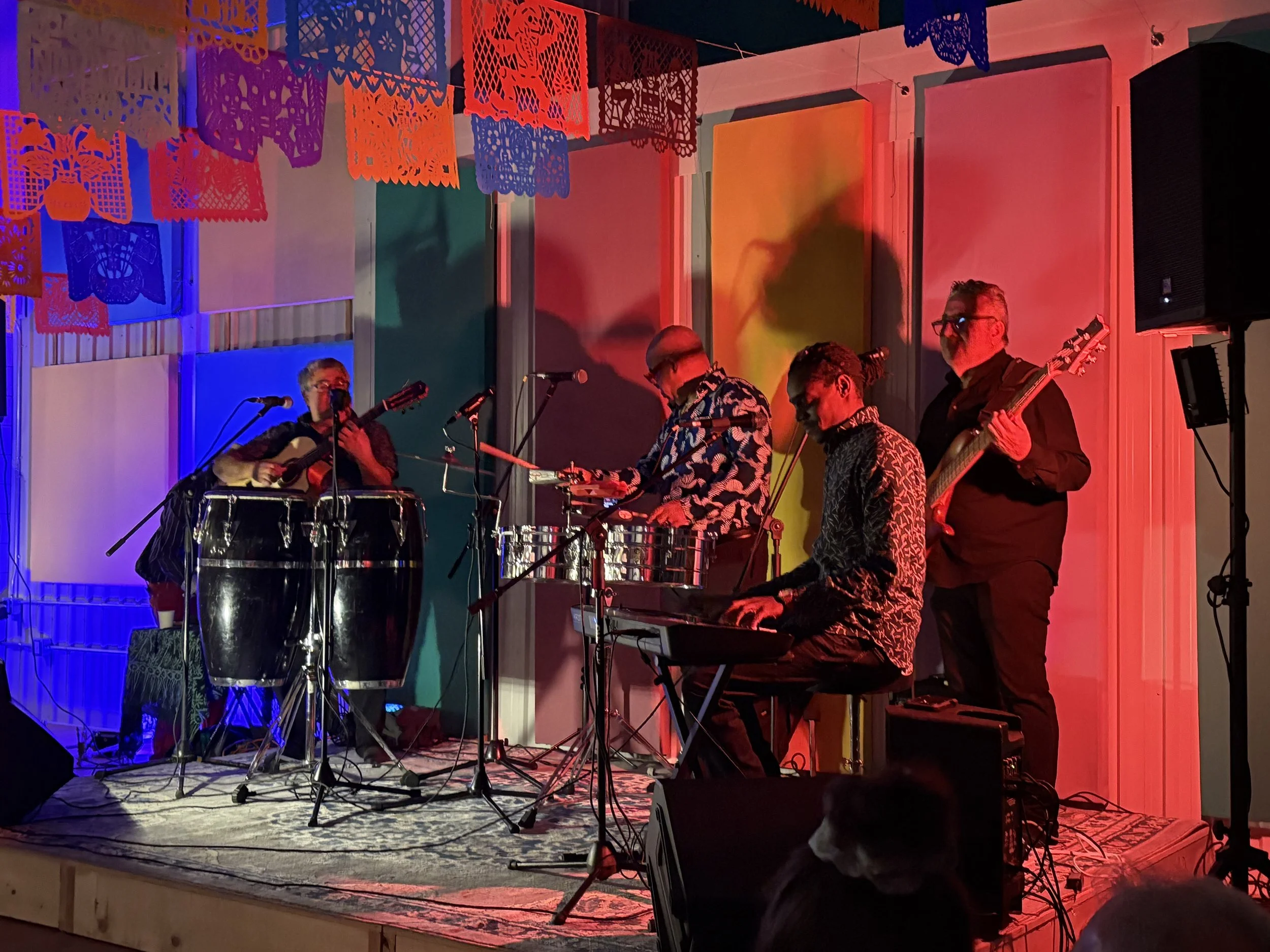A band of five musicians performing on stage with colorful lighting and papel picado decorations overhead.