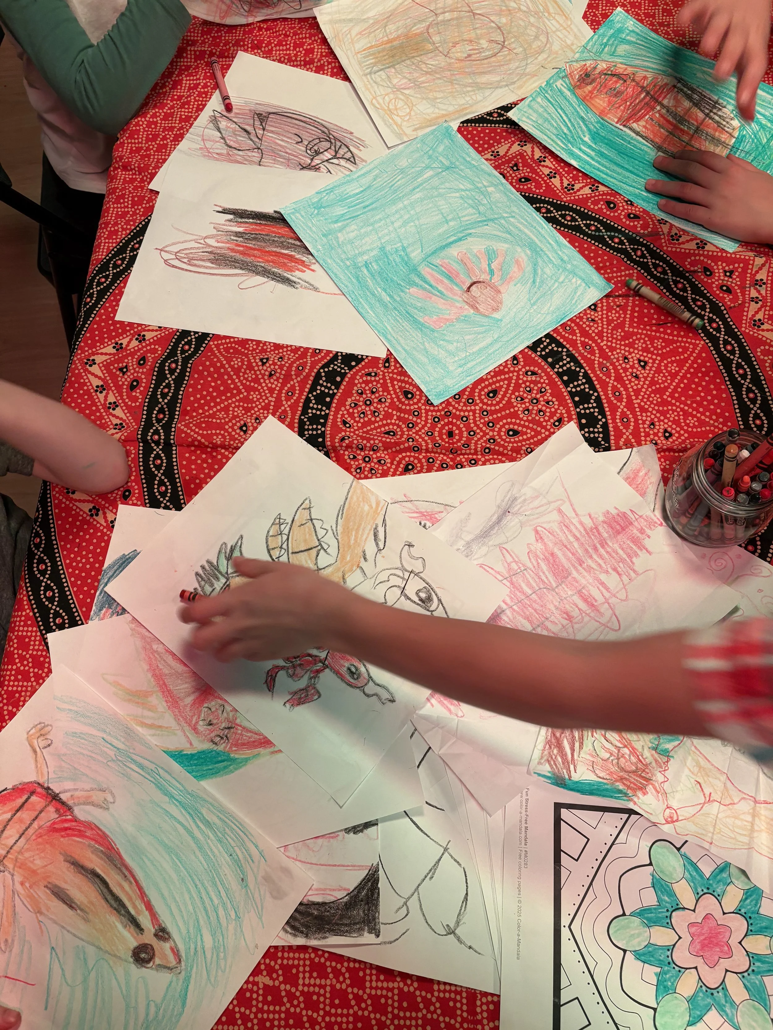 Children's drawings and coloring papers scattered on a red patterned tablecloth, with some children reaching for or holding the papers.