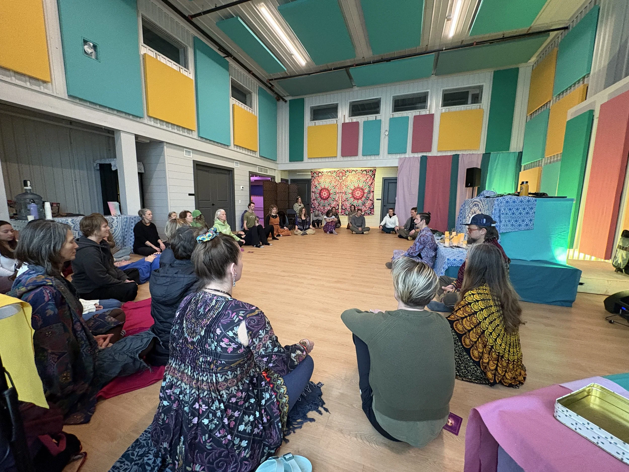 A group of people sitting on the floor in a circle in a colorful, decorated room, attending a meditation or mindfulness session.