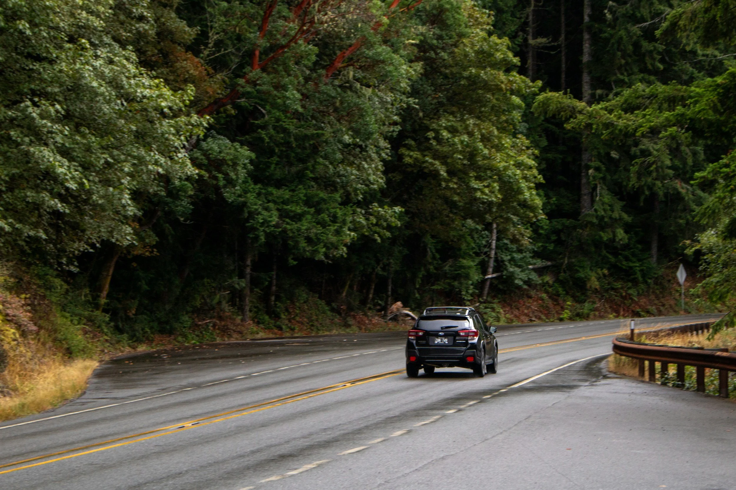 A black car driving on a winding mountain road surrounded by dense green trees.