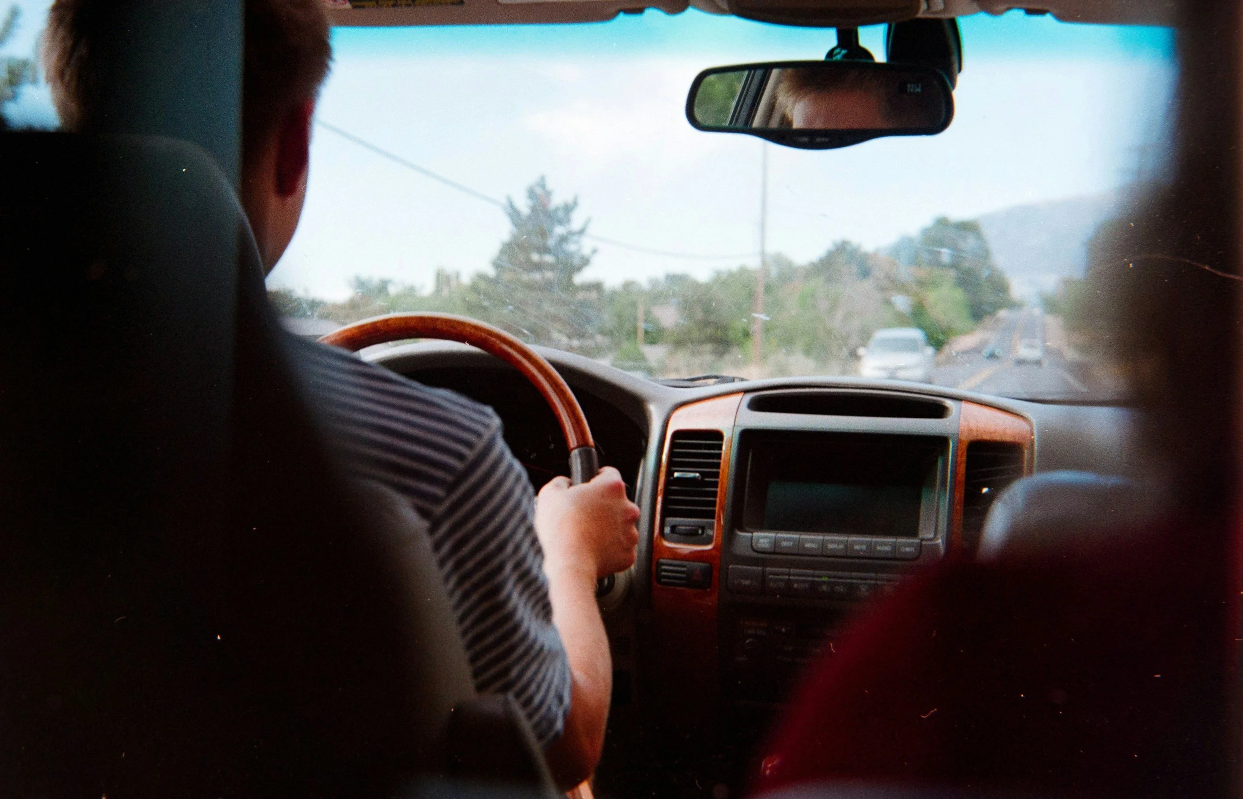 Inside a car, a person with short hair is driving on a road surrounded by trees and vehicles, viewed from the backseat through the windshield.