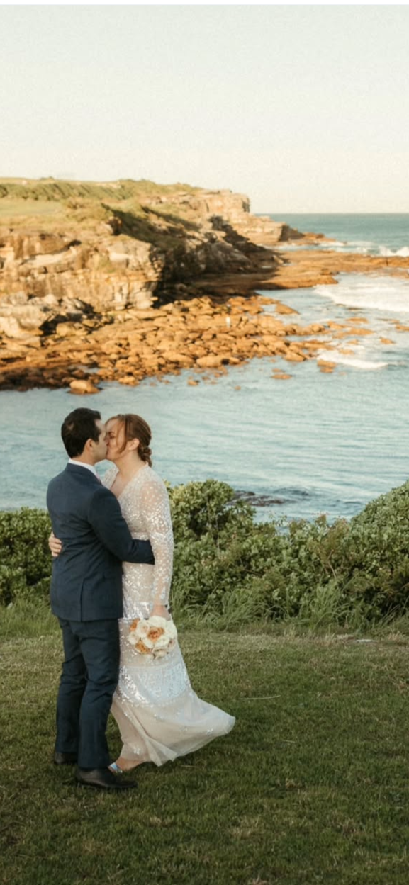 A newlywed couple sharing a kiss outdoors on a grassy area near the coast, with rocky cliffs and the ocean in the background.