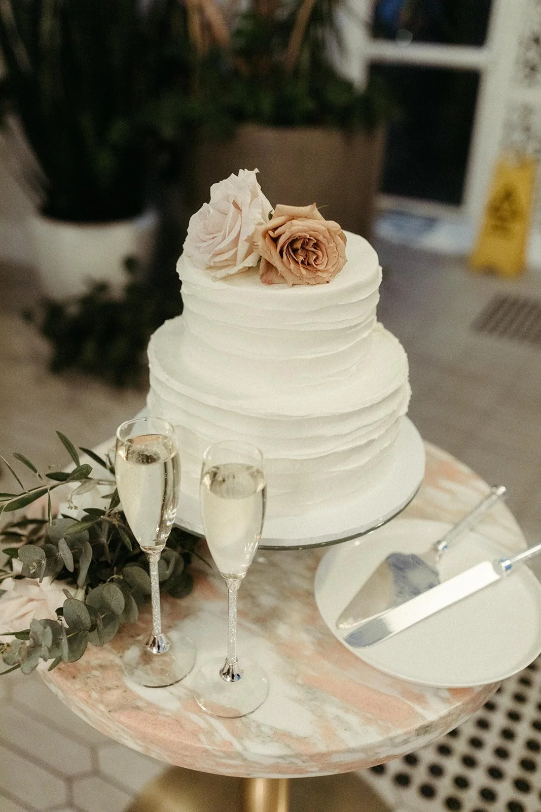 A layered white wedding cake decorated with toffee coloured roses on top, accompanied by two glasses of champagne, greenery, and cake serving utensils on a round marble table.