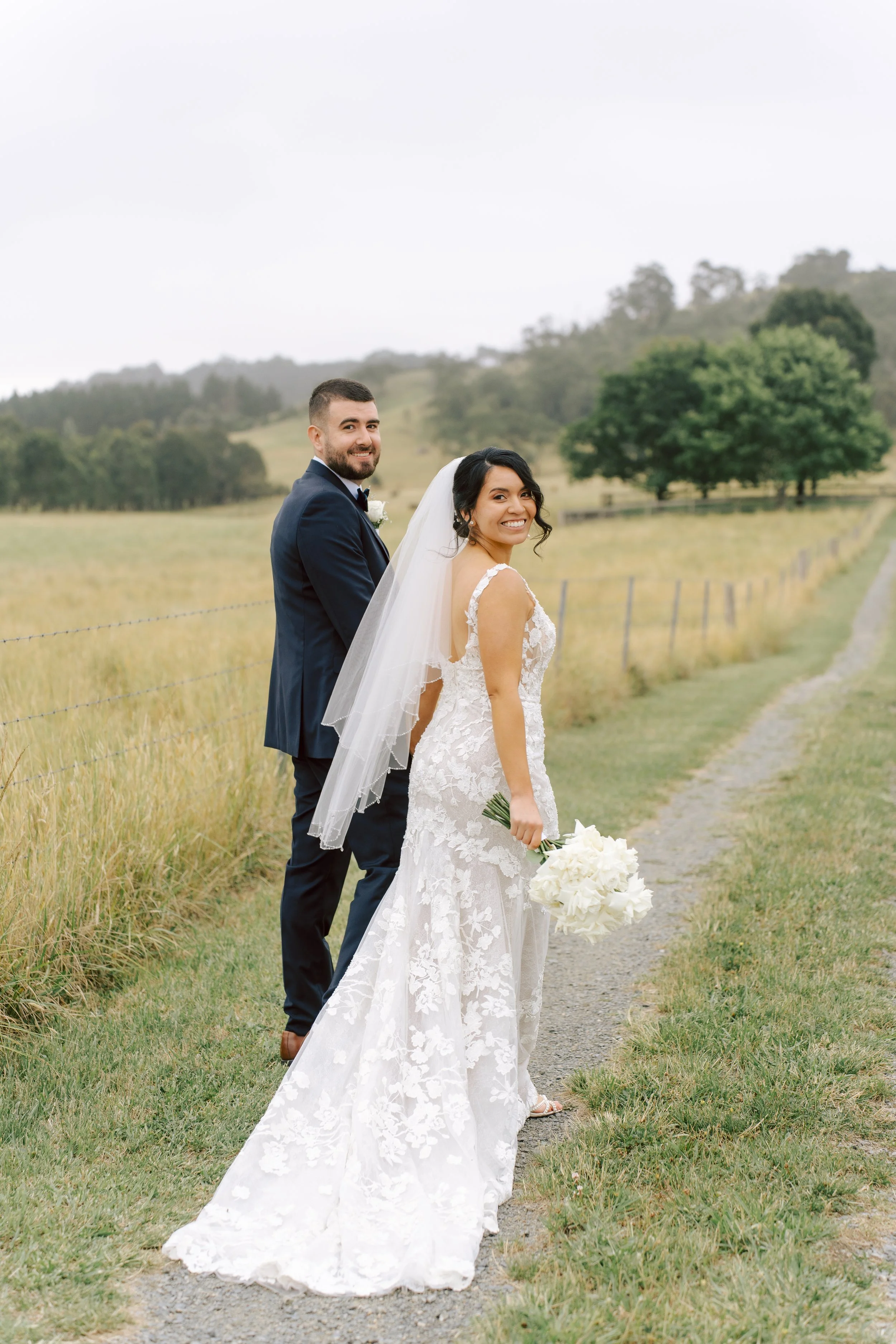 A bride and groom stand outdoors on a gravel path, smiling at the camera, with a scenic countryside background of rolling hills, trees, and grass. The bride is wearing a white lace wedding gown and holding a bouquet of white flowers, while the groom is dressed in a navy blue suit with a white shirt and bow tie.