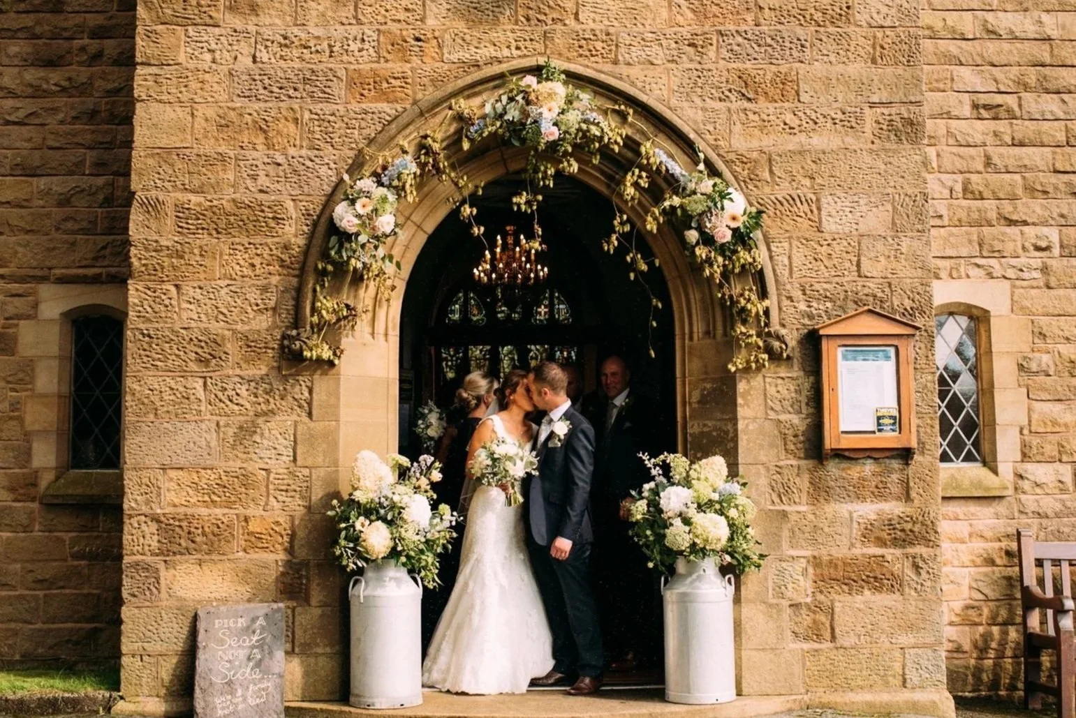 A bride and groom kissing under an arched stone church entrance decorated with flowers, with wedding guests in the background.