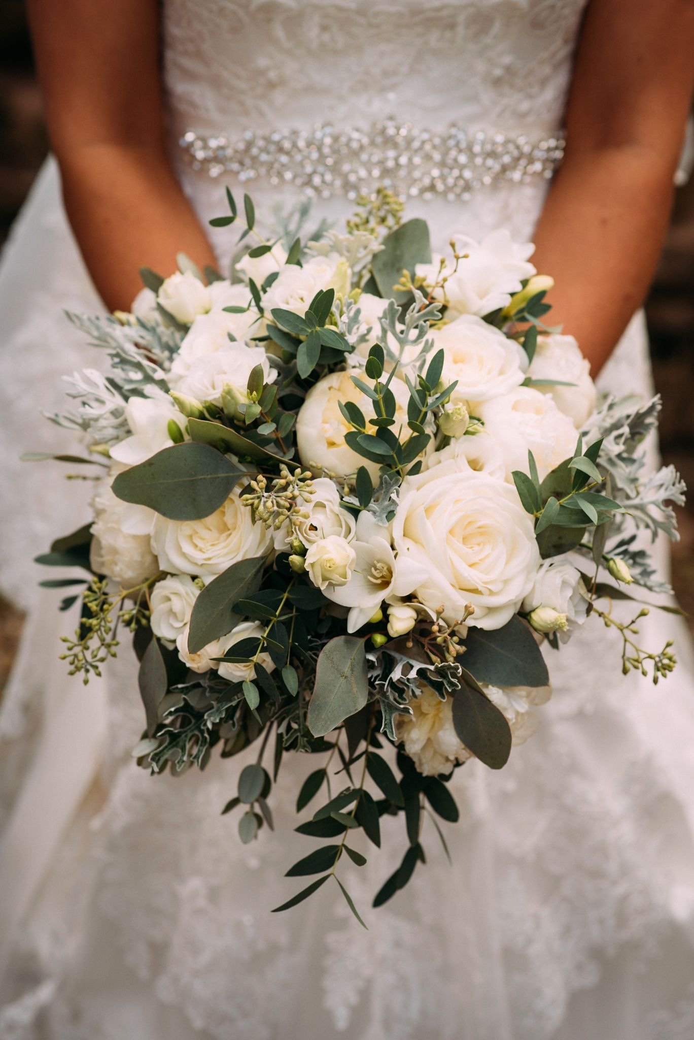 Close-up of a bride holding a bouquet of white roses, green eucalyptus, and other greenery, with the lace and beaded bodice of her wedding dress in the background.