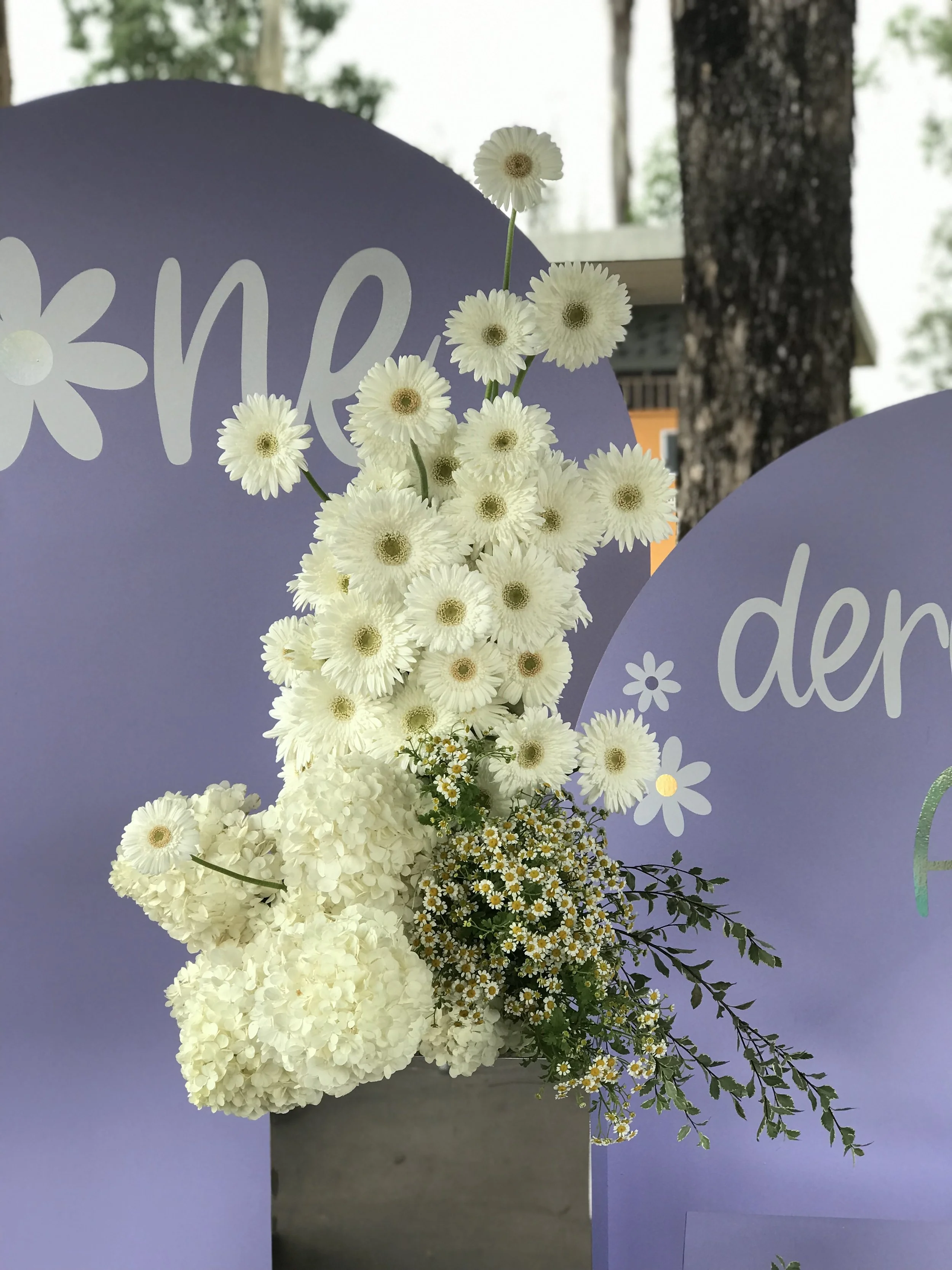 A floral arrangement with white daisies, hydrangeas, and other small flowers in a concrete vase, set against lavender signs with white floral designs.