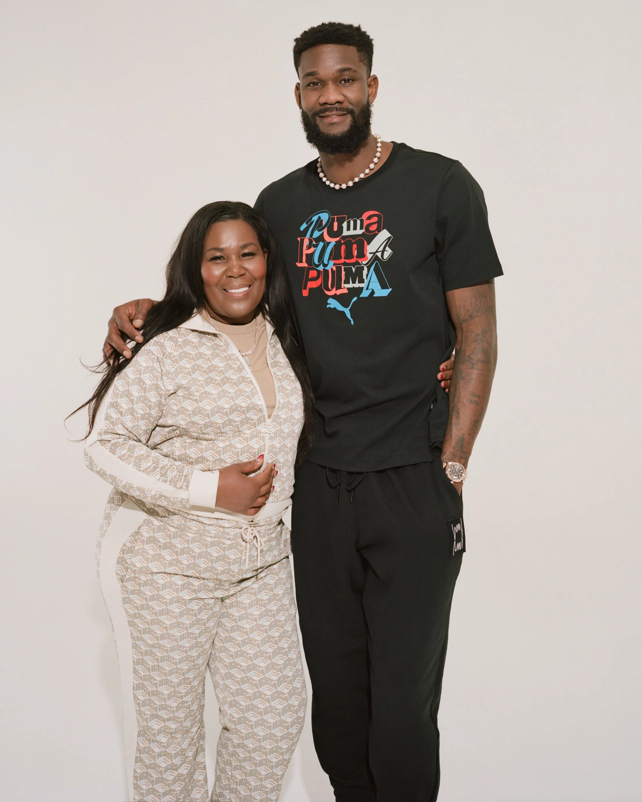 A woman and a tall man pose together in front of a plain white wall, smiling. The woman wears a beige patterned tracksuit, while the man is in black Puma t-shirt, black pants, and a pearl necklace.