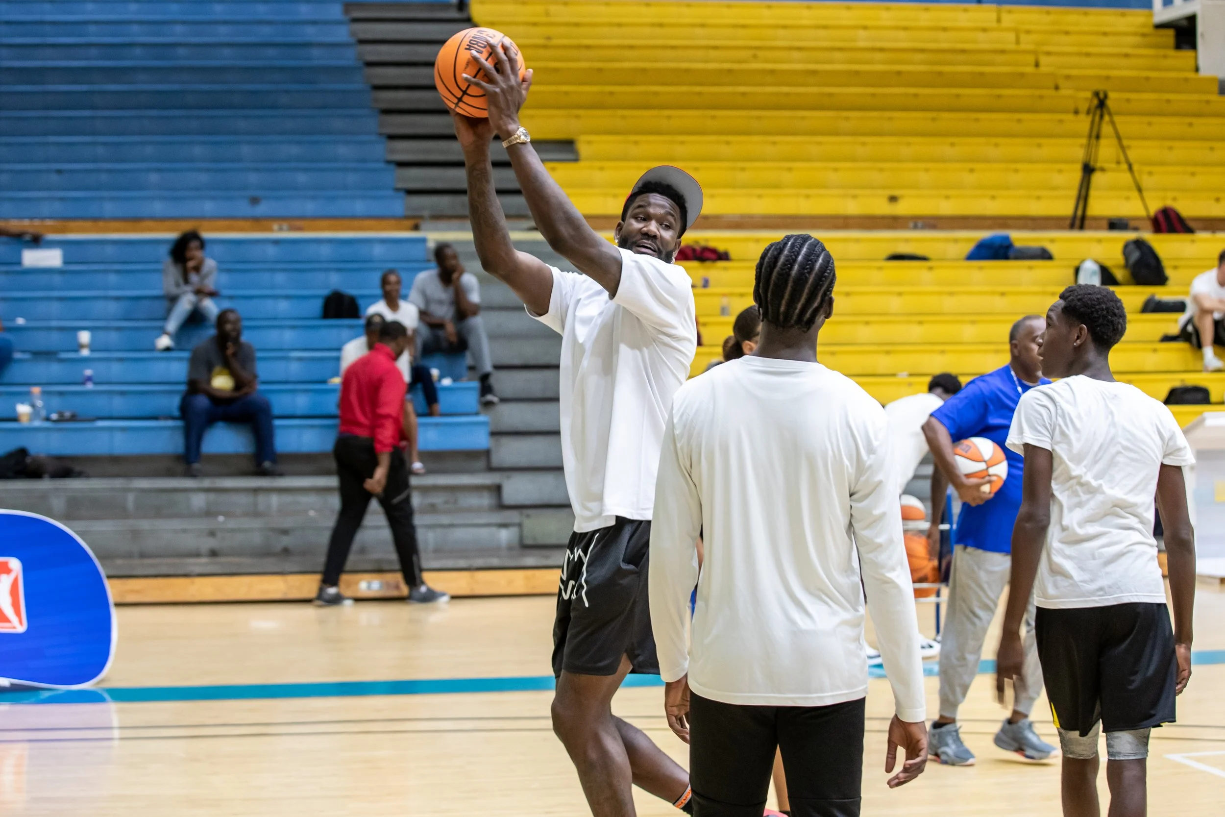 A group of young men playing basketball on an indoor court, with one man preparing to shoot the ball. Some spectators are sitting on bleachers in the background.