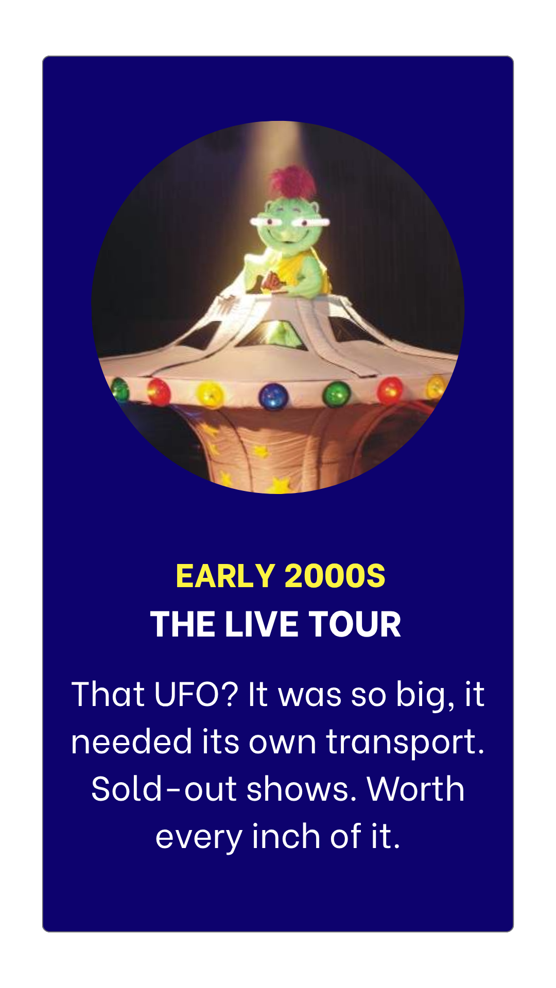 A puppet of a green alien with red hair sitting in a small UFO prop, decorated with colorful lights, on stage during a concert or performance.