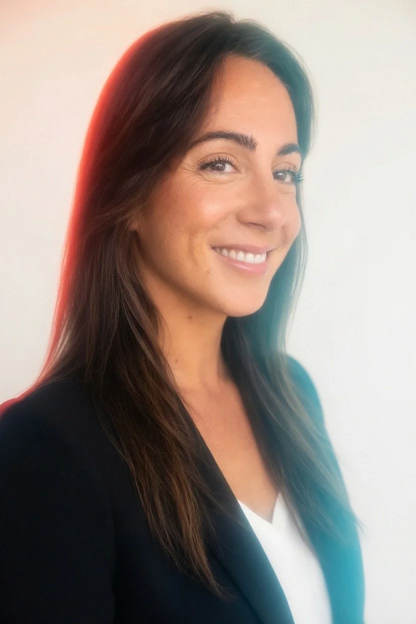 A woman with long dark hair, smiling, wearing a black blazer over a white top, against a plain light-colored background.