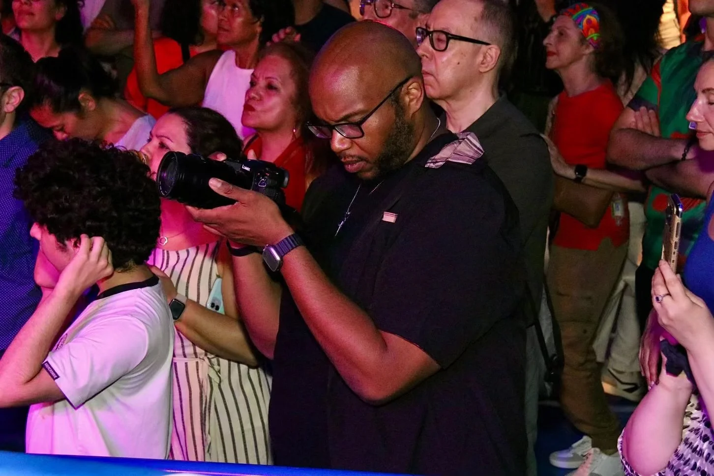 A man taking a photograph with a camera in a crowded indoor setting with diverse people, some with their arms crossed or talking, illuminated by pink and purple lighting.