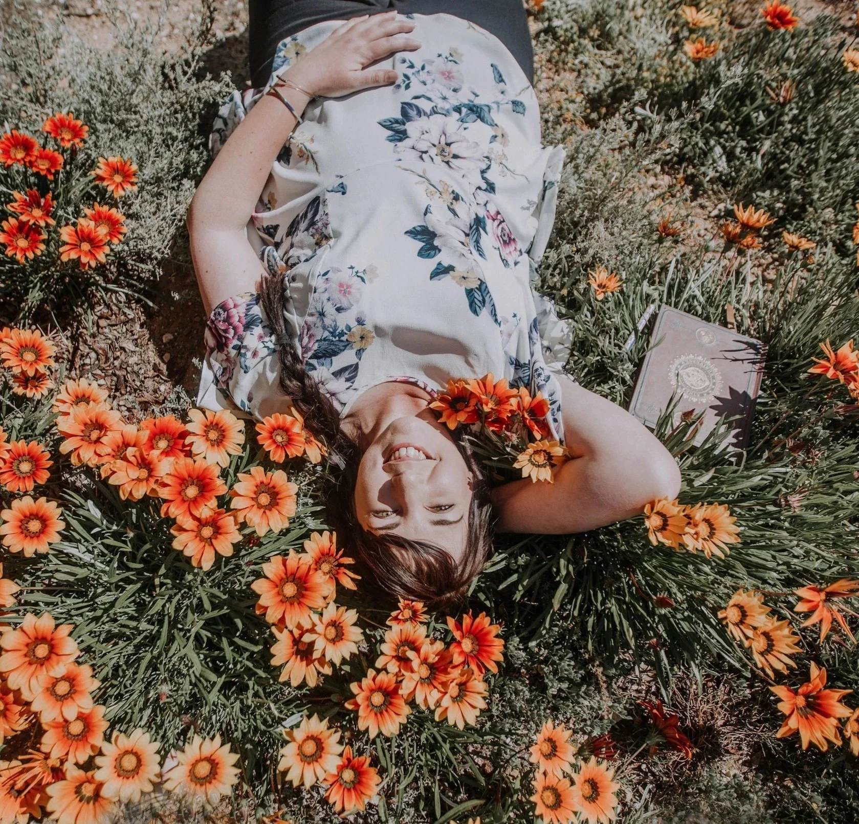 A smiling woman with brown hair lying on her back in a bed of vibrant orange and yellow flowers, wearing a floral dress and bracelets, with a plant and a book nearby.