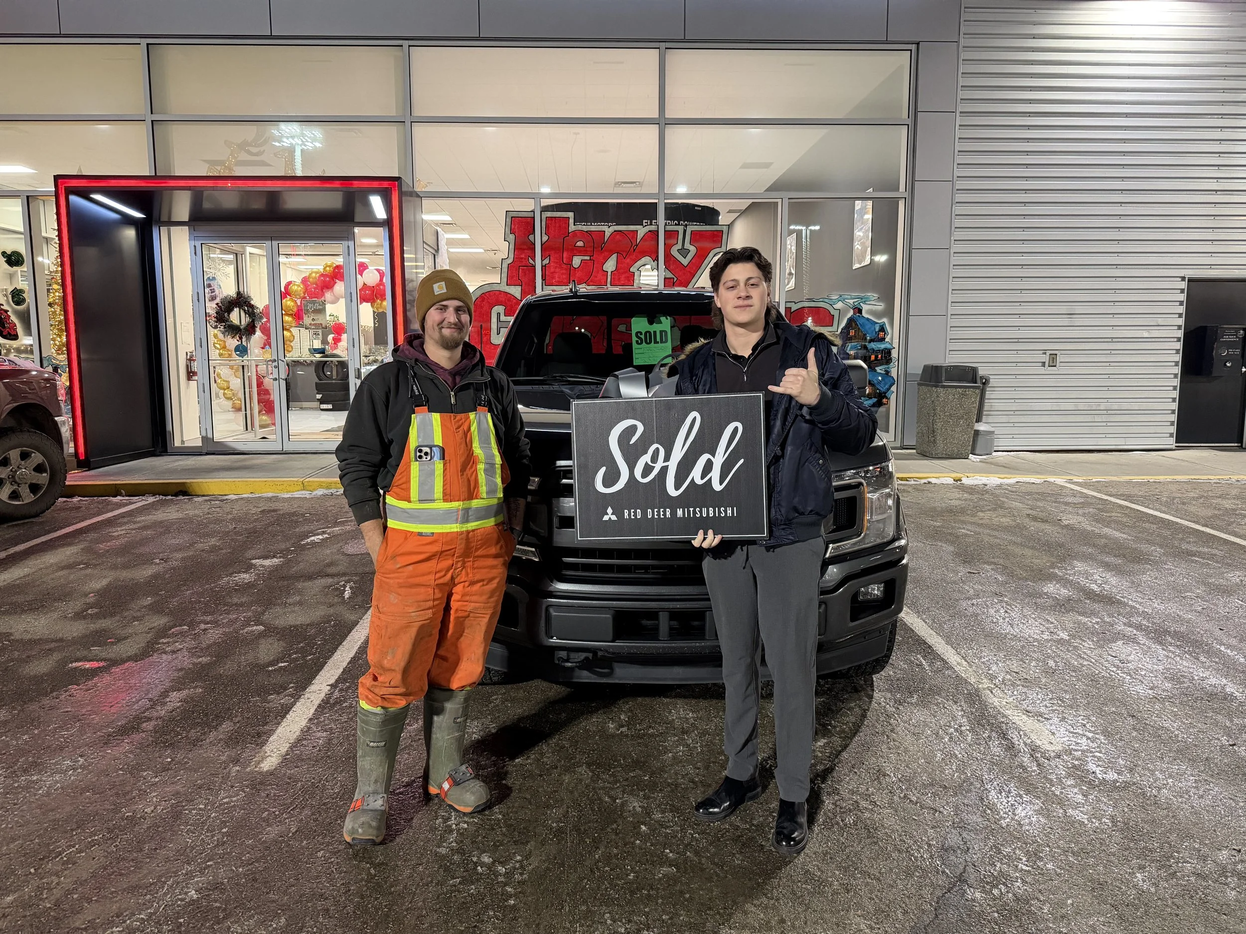 Two men standing in front of a black pickup truck outside a building, with one holding a 'Sold' sign. The man on the left is dressed in work clothes, including orange pants, a high-visibility vest, and rubber boots. The man on the right is wearing a dark jacket and pants, and is making a shaka sign with his hand.