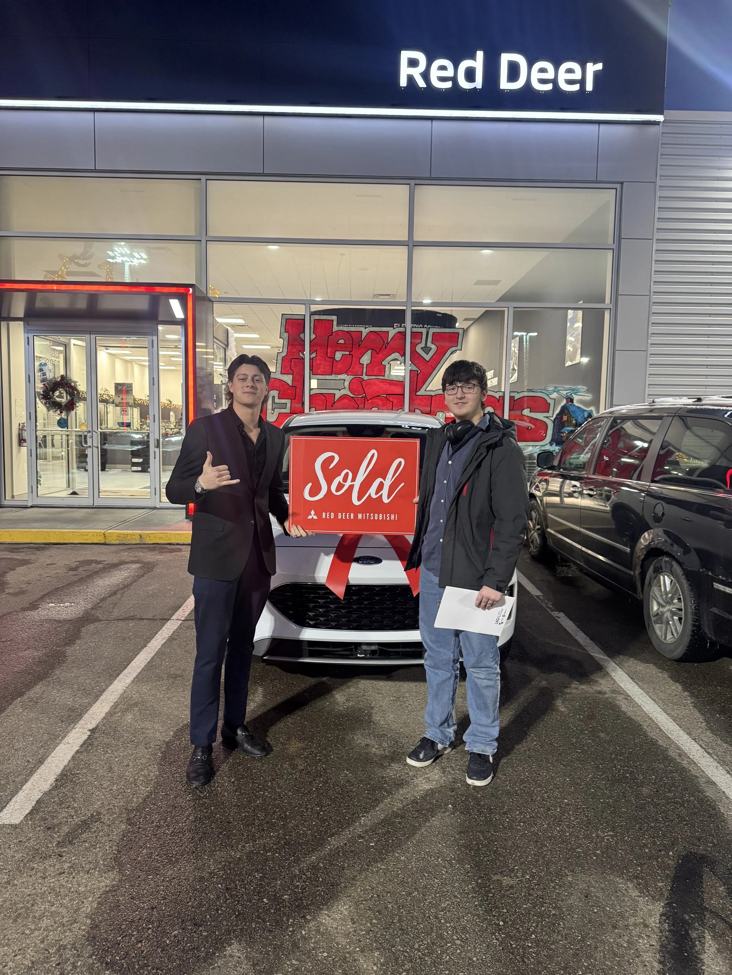 Two young men standing in front of a white car with a red 'Sold' sign in a dealership parking lot at night, outside a building with a sign that reads 'Red Deer' and a glass door entrance decorated for Christmas.