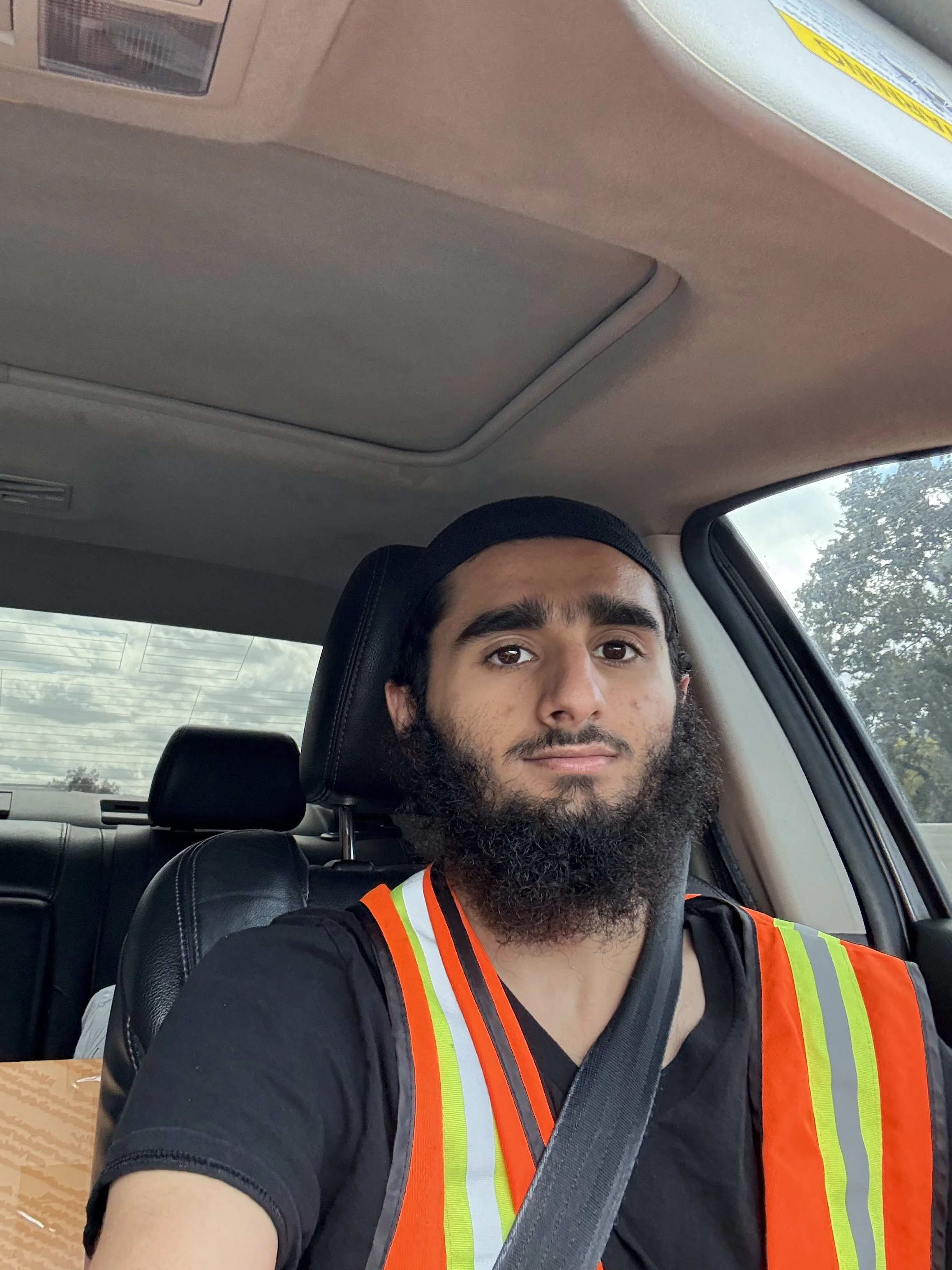 A man with a beard and dark hair wearing a black shirt and safety vest taking a selfie inside a vehicle.