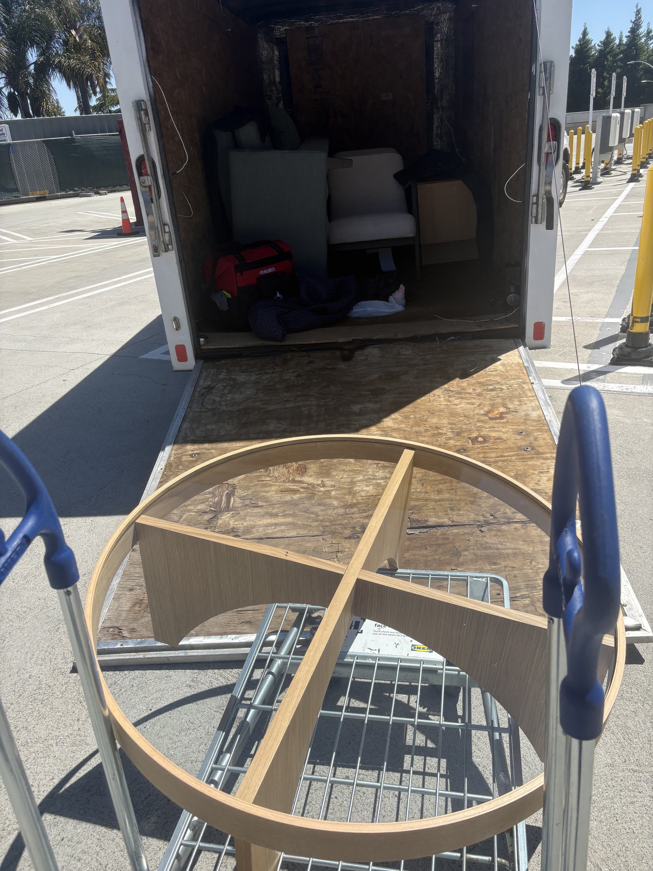 View from a shopping cart looking into the back of a moving truck with furniture inside, including a round wooden tabletop, a light-colored armchair, and a gray upholstered chair, in a parking lot outside.