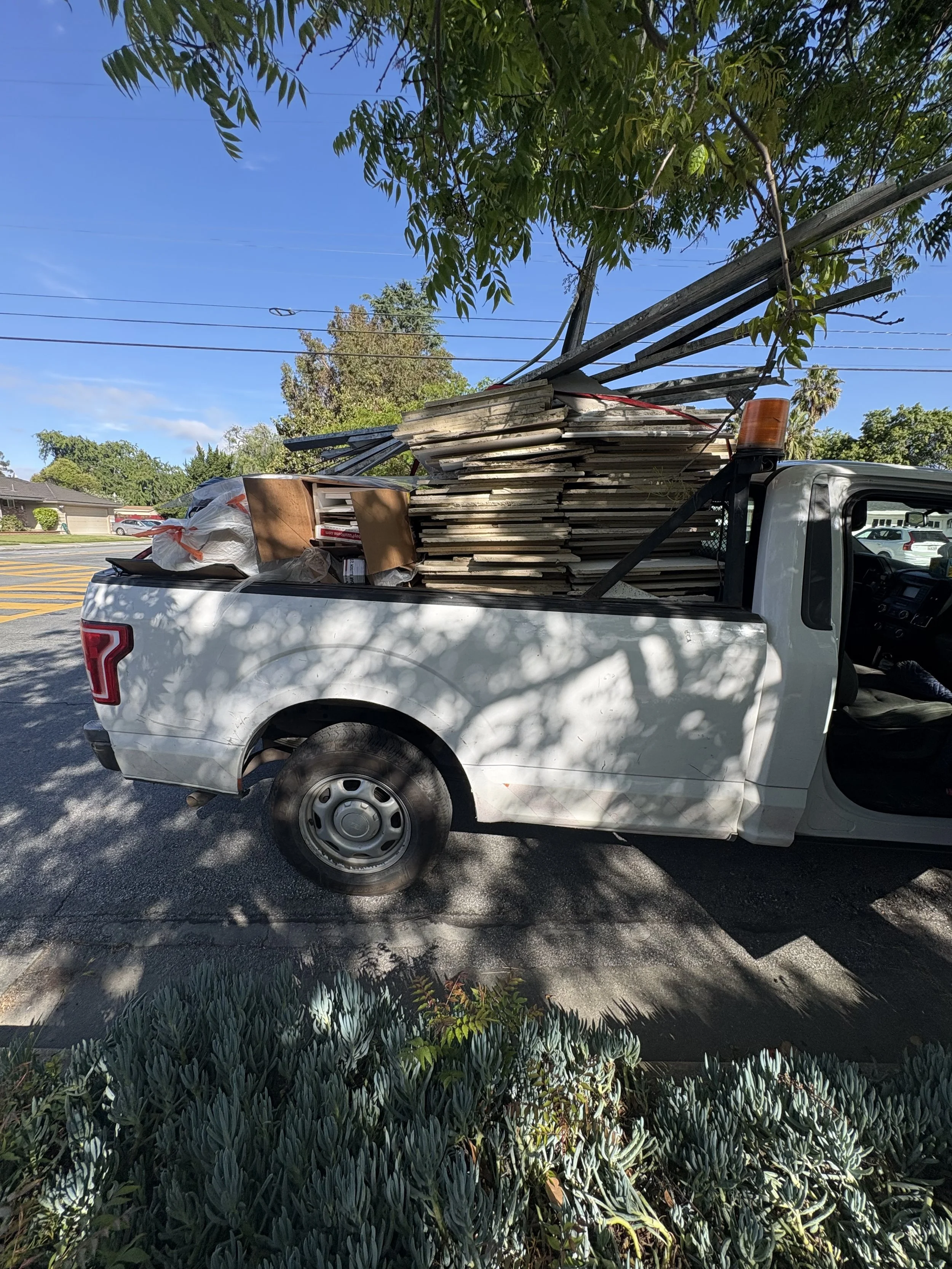White pickup truck loaded with wooden planks, boxes, and other construction materials parked on a street near green plants and trees.