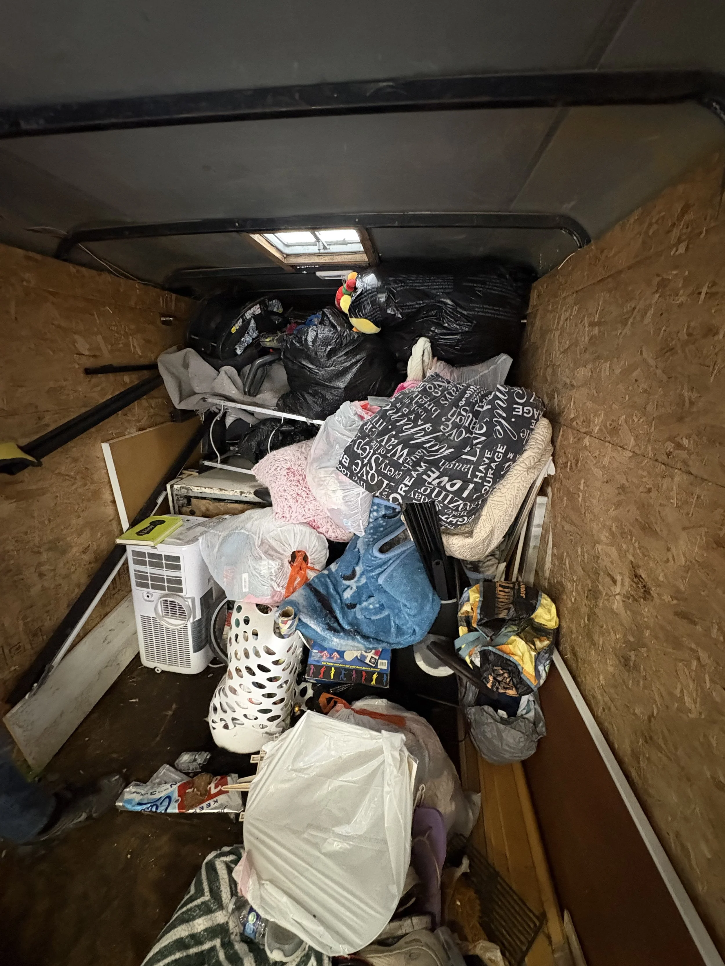 A cluttered room with various items including a small space heater, a laundry basket, bags, clothes, and miscellaneous objects scattered across the floor and on a desk.