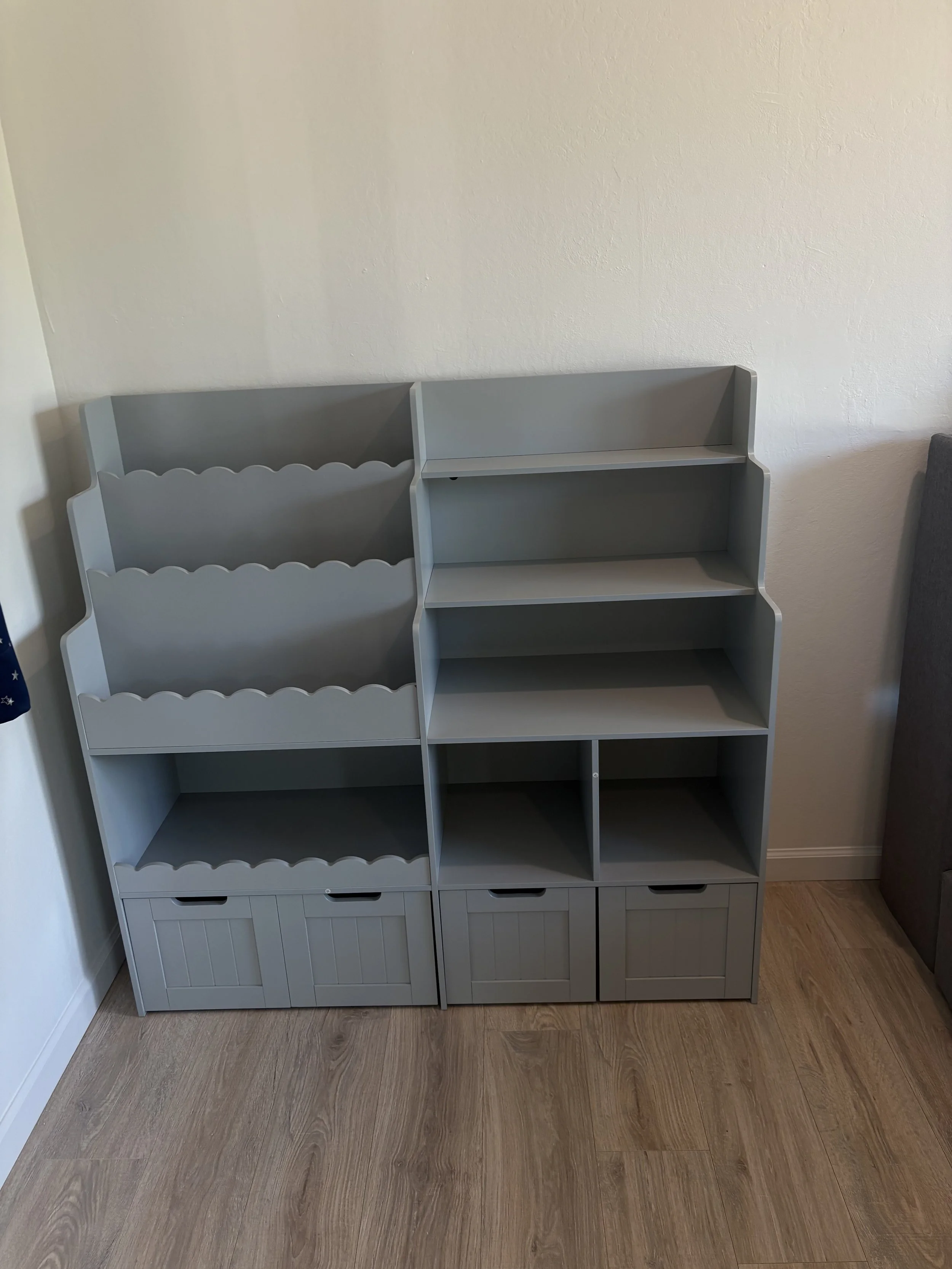 Gray bookshelf with three shelves and three storage drawers at the bottom, positioned against a white wall on wood flooring.