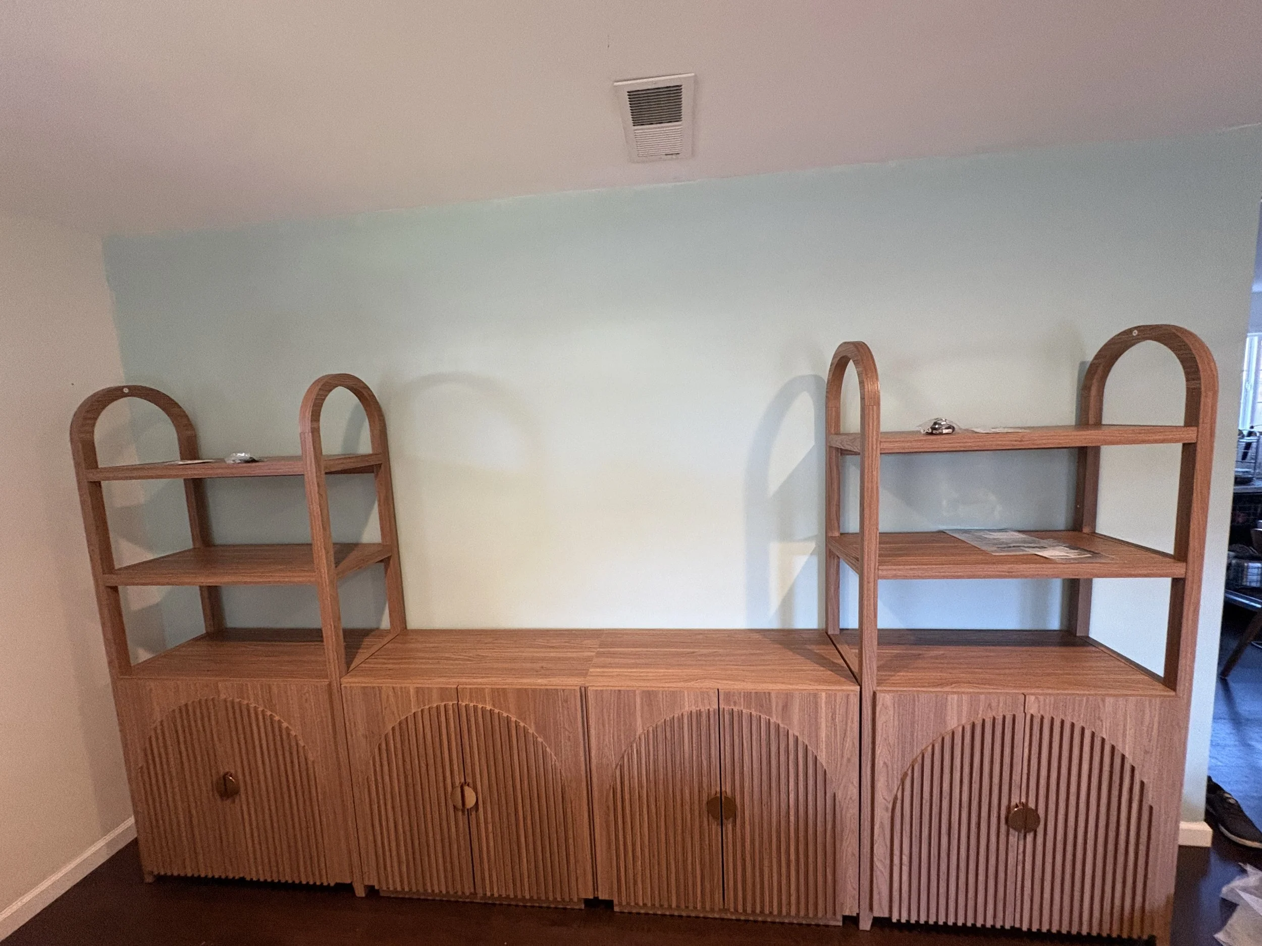 Empty wooden shelving unit with three arched cabinet doors at the bottom and two shelves on top, settled against a light green wall. A small silver object is on the right shelf.
