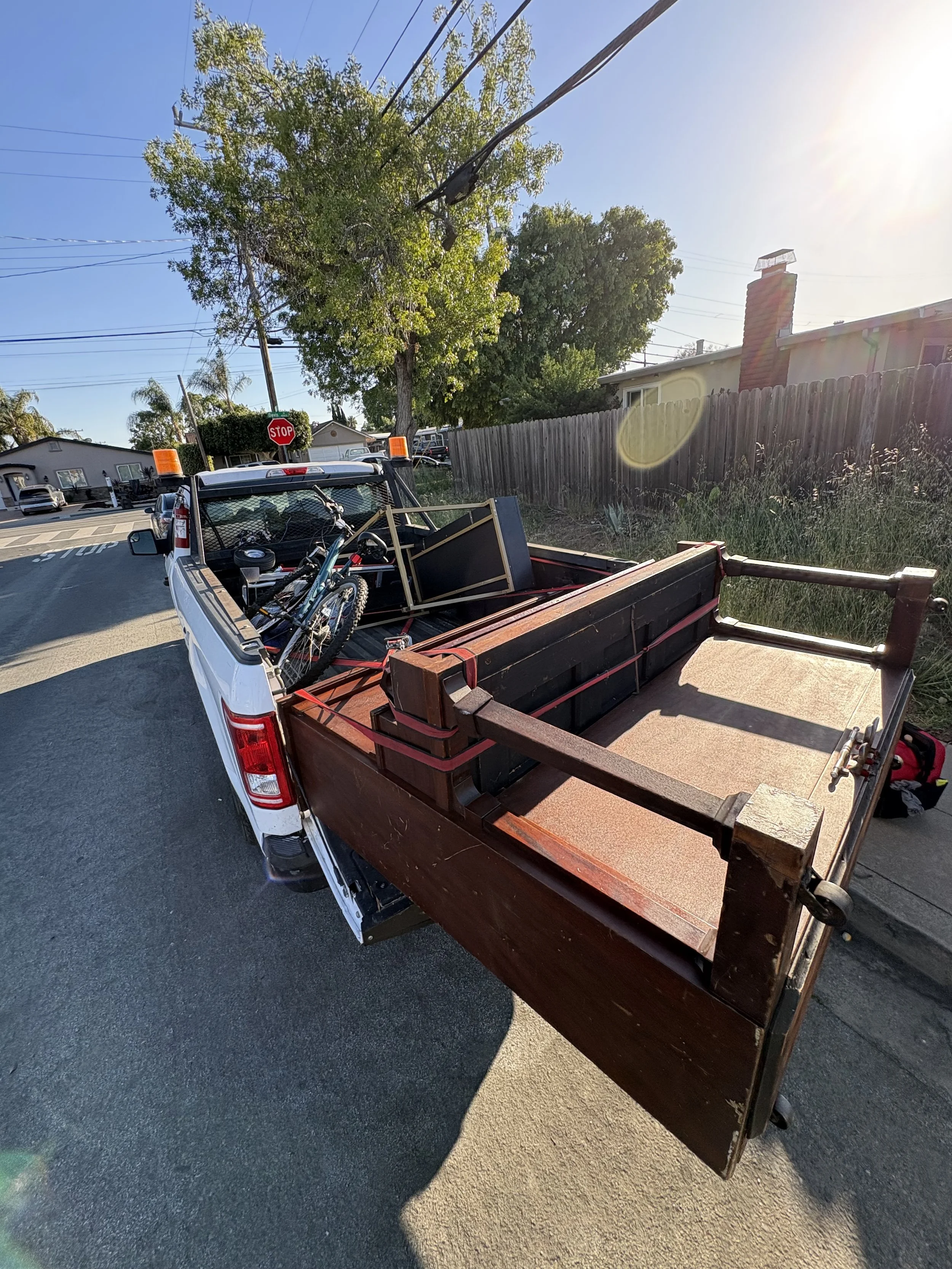 A white pickup truck parked on the street with a wooden trailer bed loaded with various tools and objects, including a bicycle. The scene is lit by sunlight, with a wooden fence, trees, and residential houses in the background.