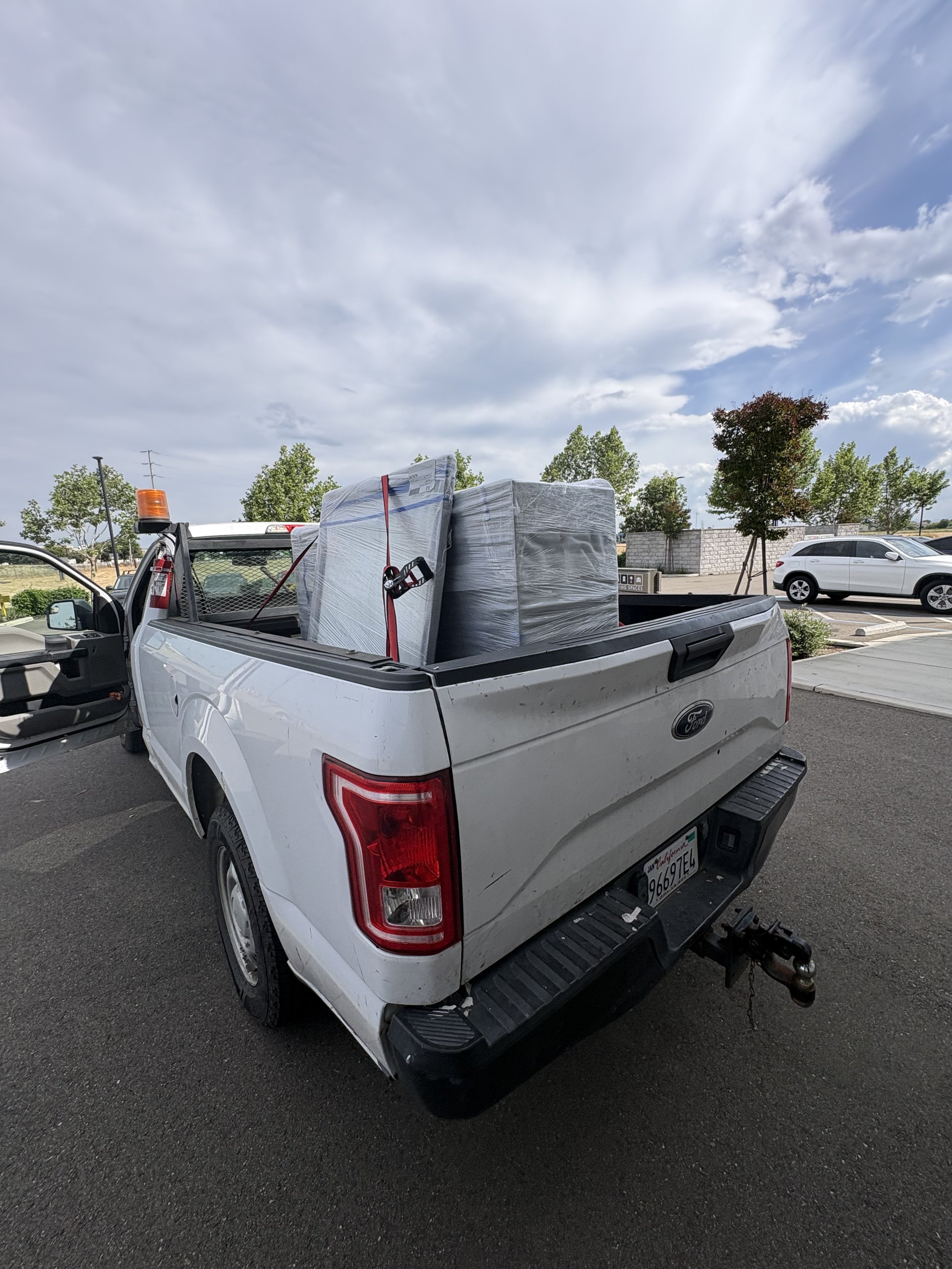 A white pickup truck parked in a parking lot, loaded with large wrapped packages in the truck bed, with trees and a cloudy sky in the background.
