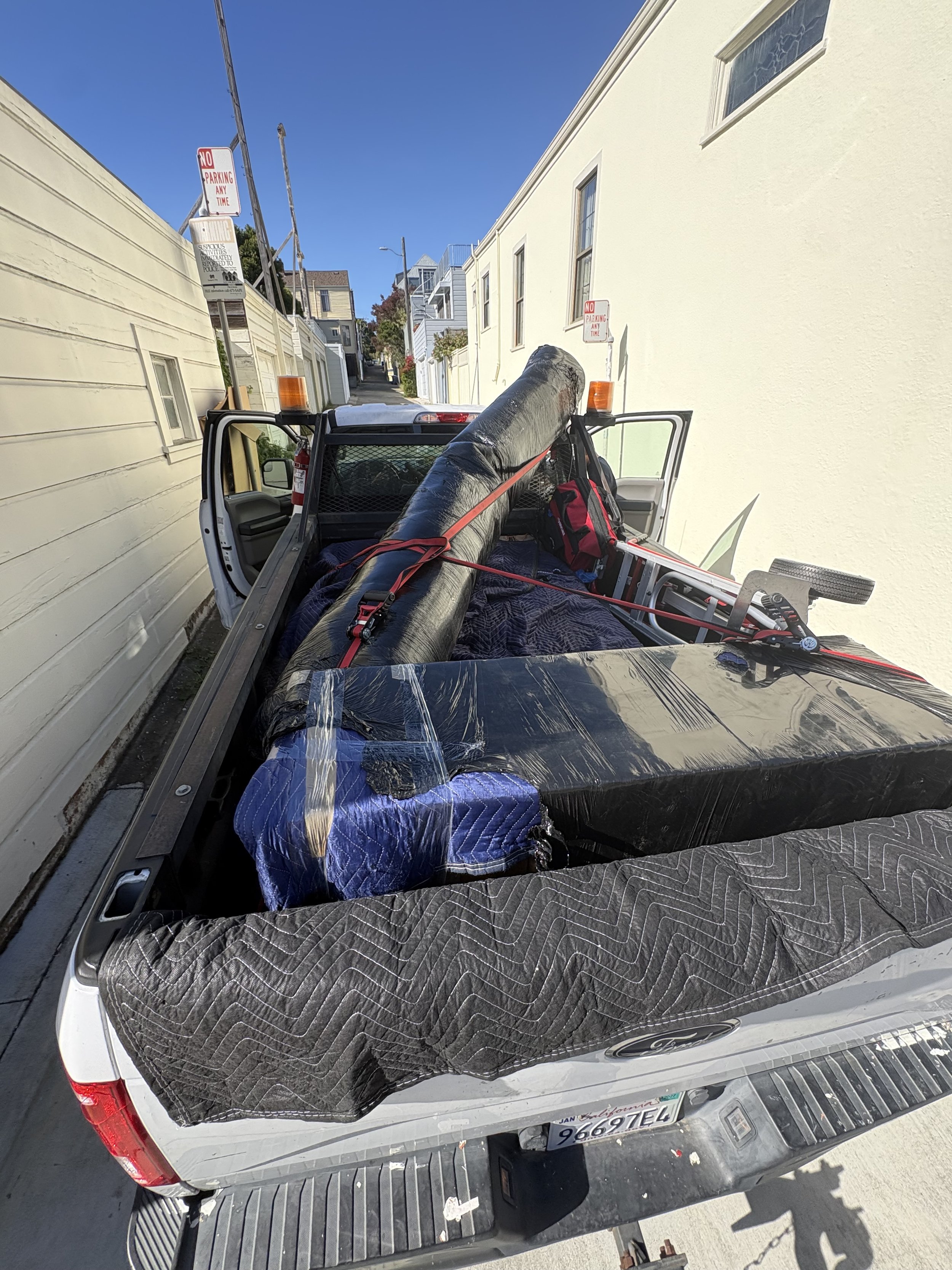 A white Ford truck with a black mat in the bed, carrying a large black pipe secured with red straps, parked on a street with residential buildings and no parking signs visible.