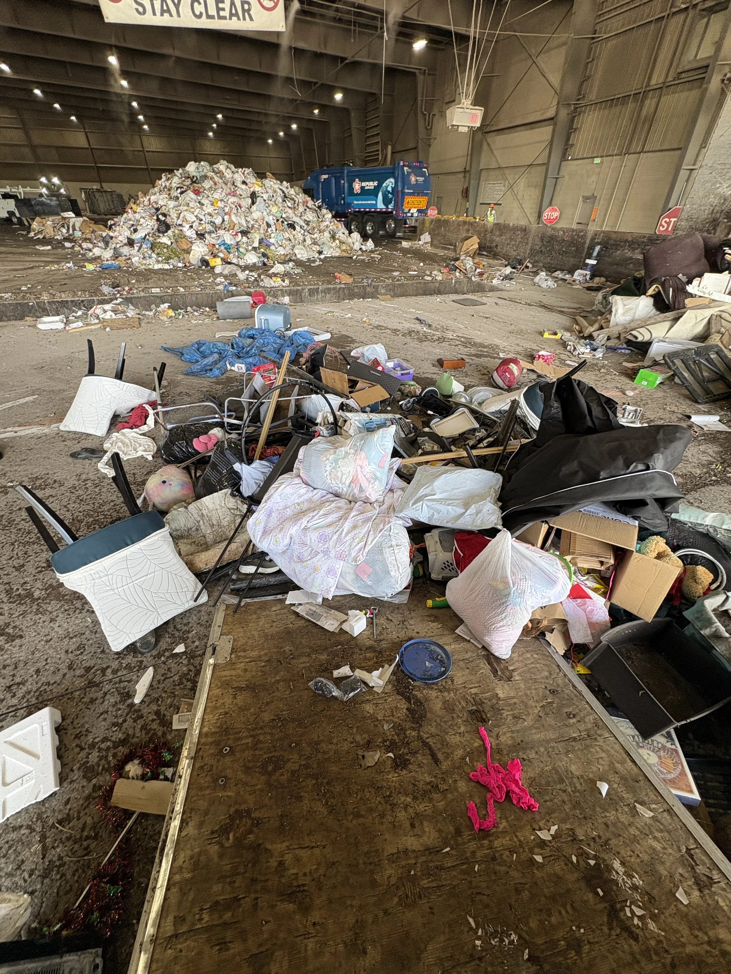 A cluttered indoor space with scattered trash, furniture, and personal belongings, including a large pile of garbage near the back and various items like chairs, boxes, and bedding on the floor.