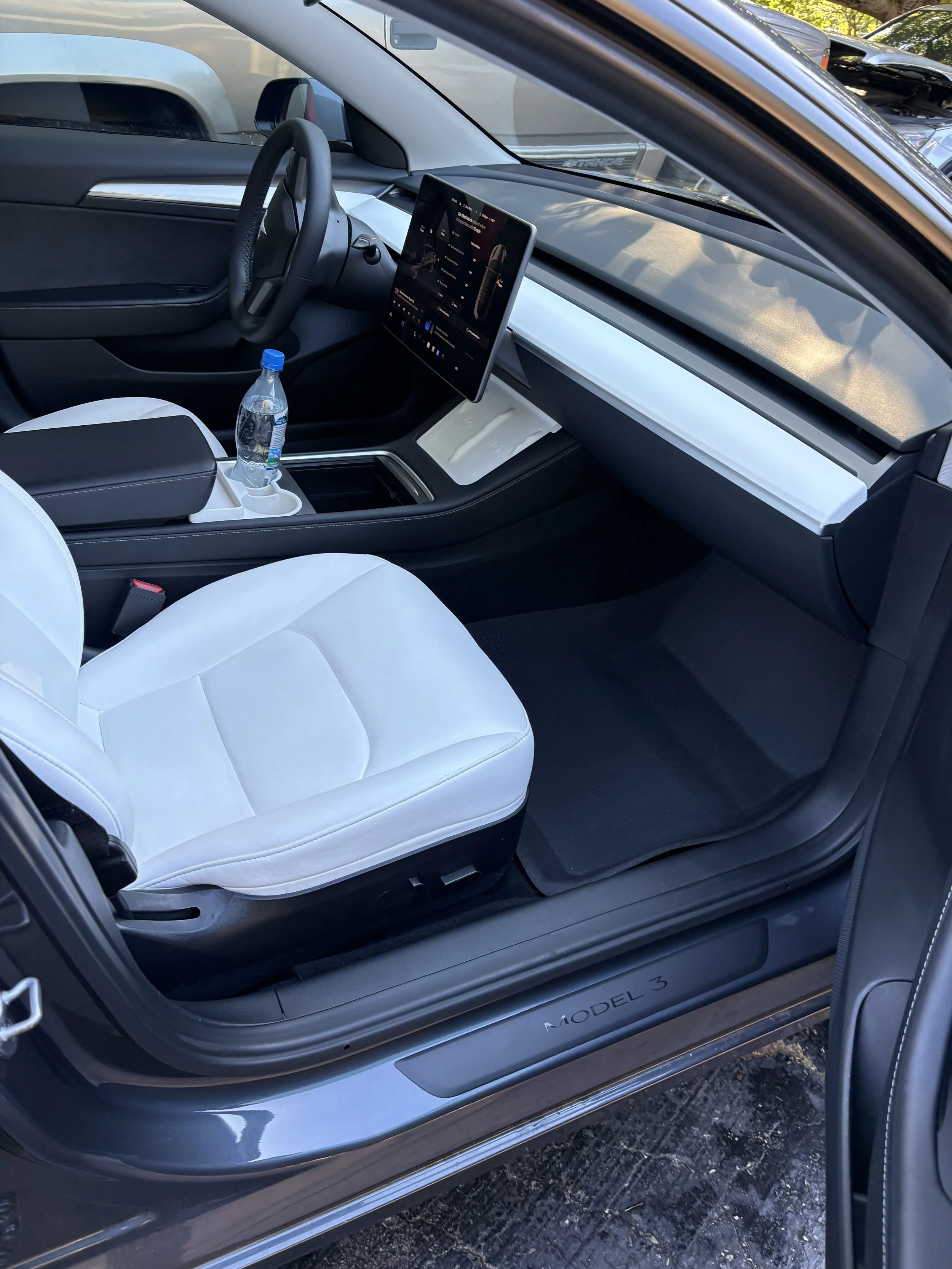 Interior of a Tesla Model 3 showing the white seat, black dashboard, steering wheel, and a large touchscreen display. There is a water bottle in the cup holder.