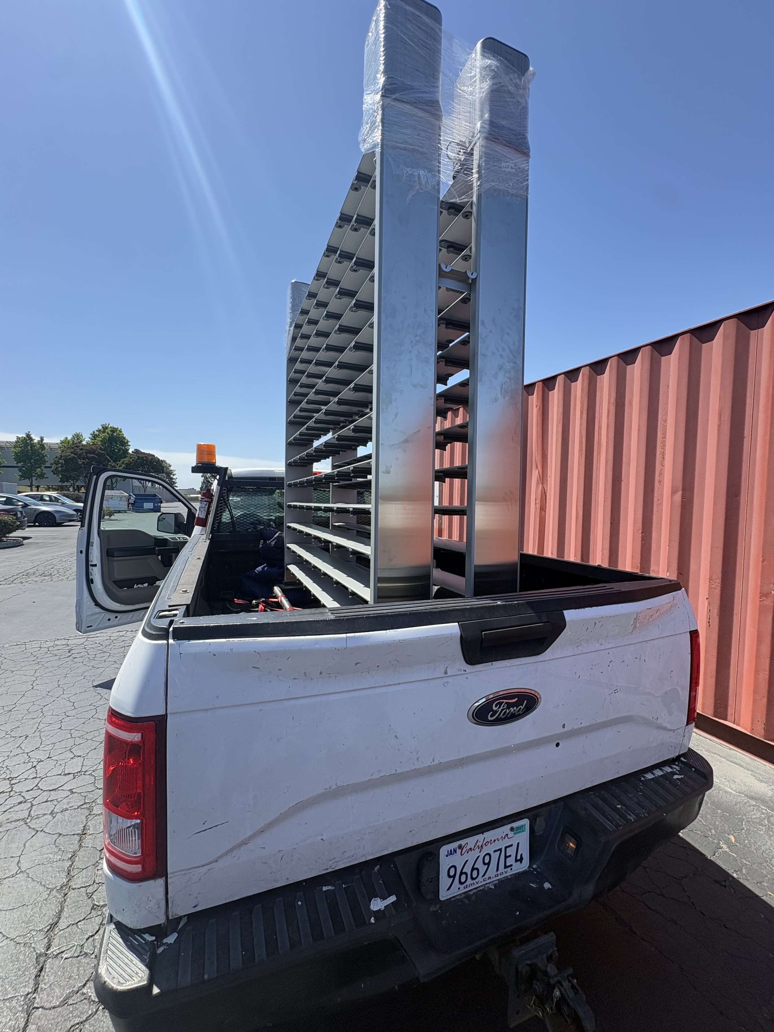 A white Ford pickup truck with a red and white California license plate, carrying two large metal racks in its bed, parked near a red fence under a clear blue sky with sunlight streaming in.