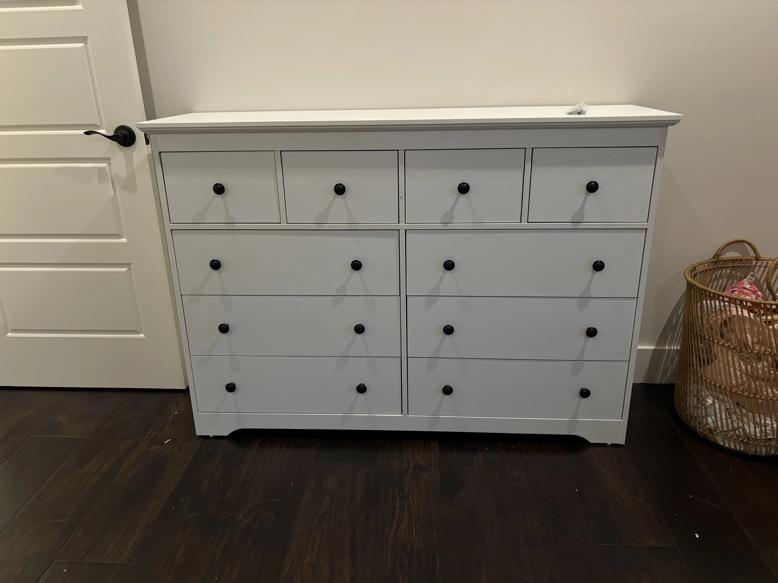 A white dresser with nine drawers and black knobs standing against a wall in a room with dark wood flooring. To the right, a wicker basket is partially visible.
