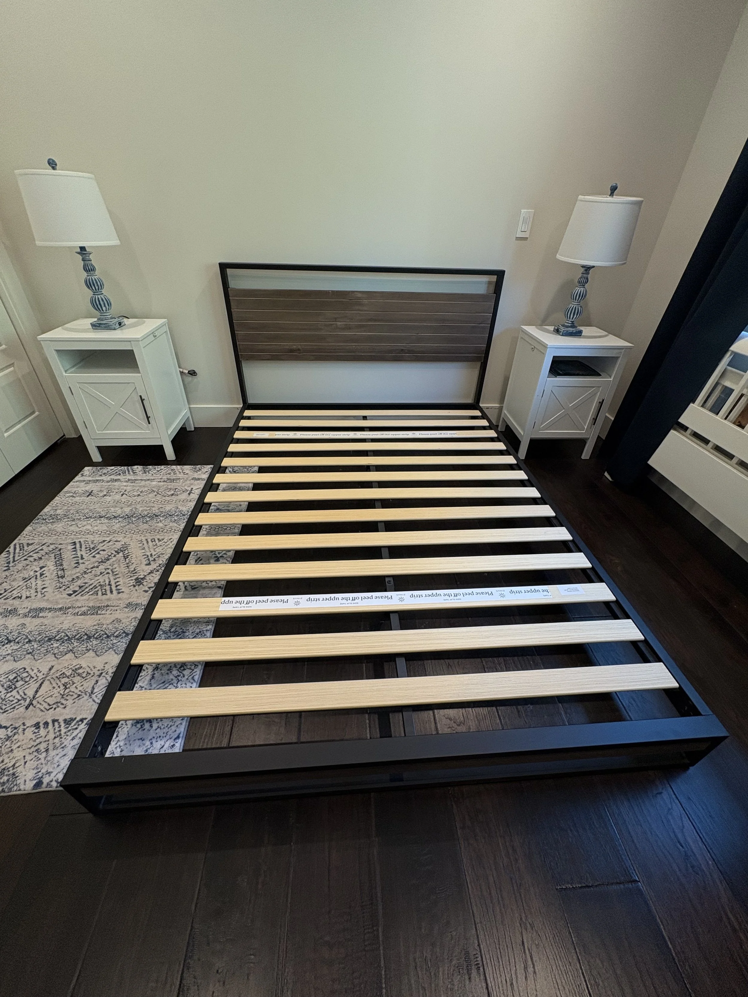 Empty bed frame with wooden slats in a bedroom, flanked by two white nightstands with lamps, on dark hardwood floor with a patterned rug nearby.