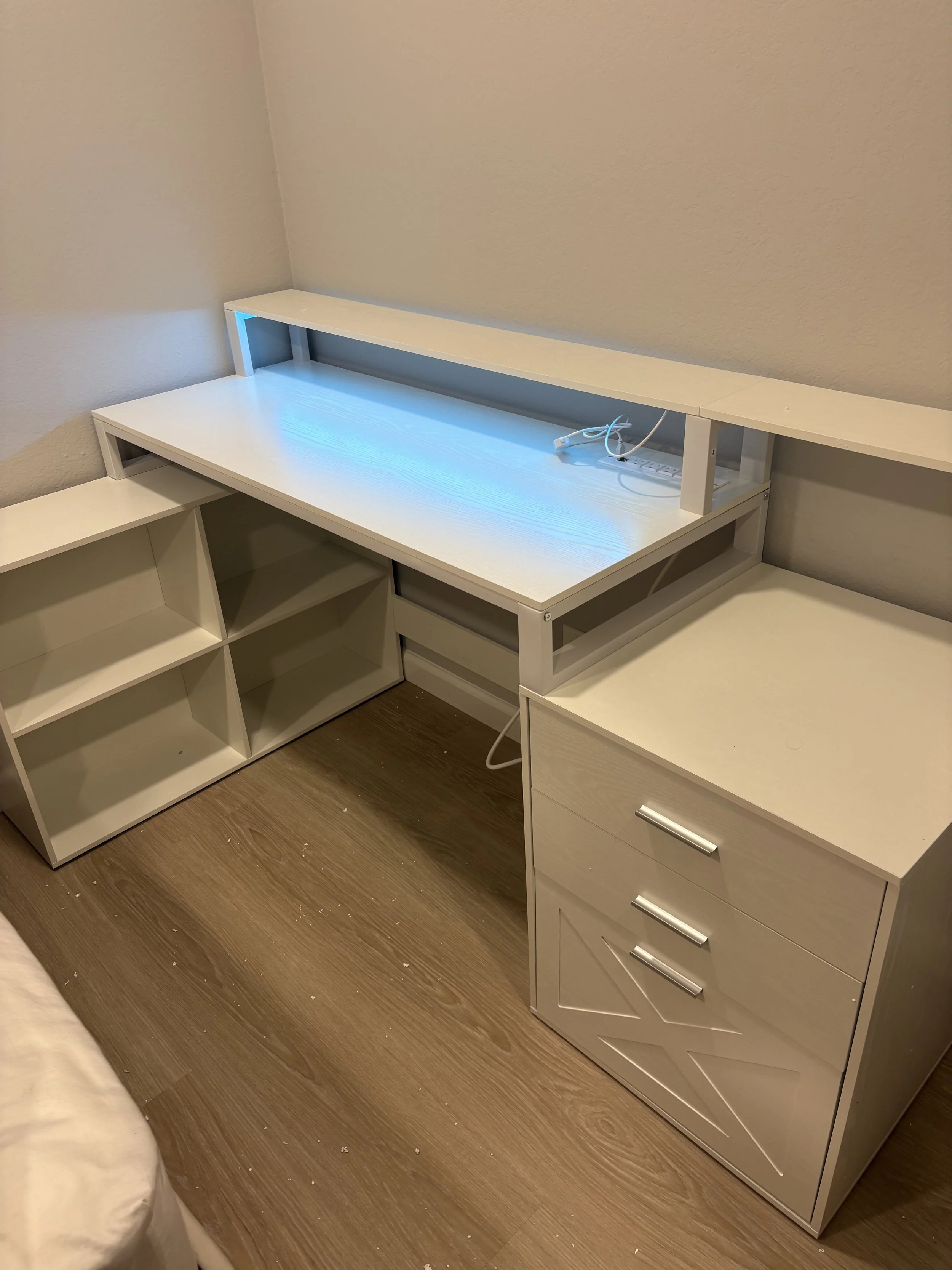 Empty white desk with a raised shelf and an electrical outlet, surrounded by white shelves and a white dresser against a beige wall, with a wooden floor.