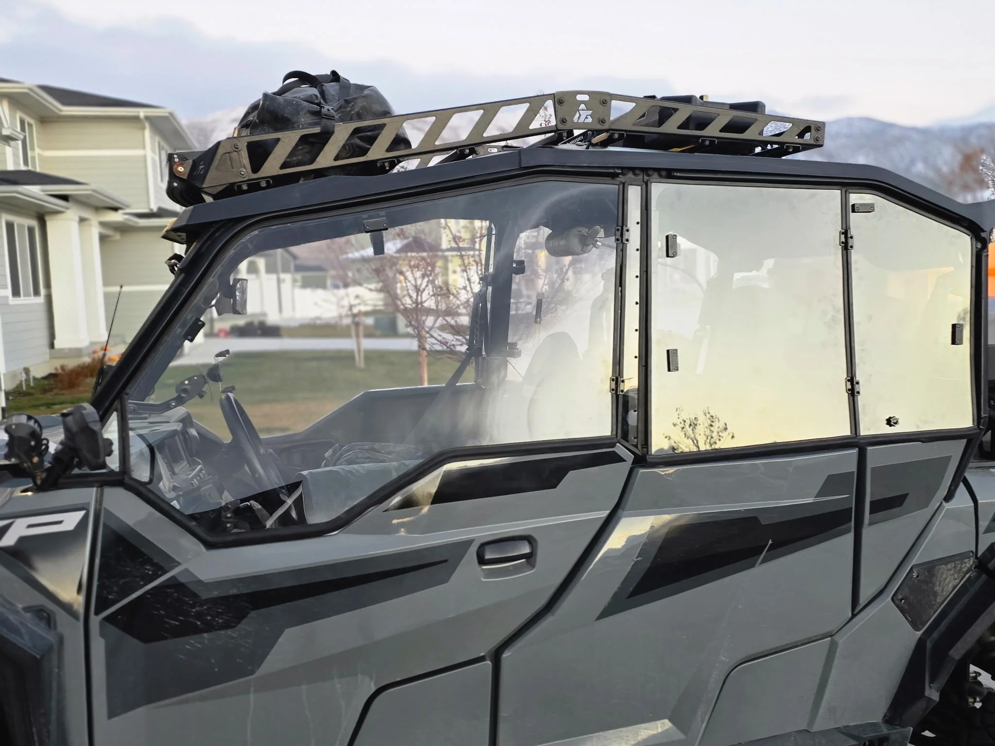 A close-up of an off-road utility vehicle with a clear plastic enclosure, black roof rack, and mounted gear, parked in a residential neighborhood with houses and mountains in the background.